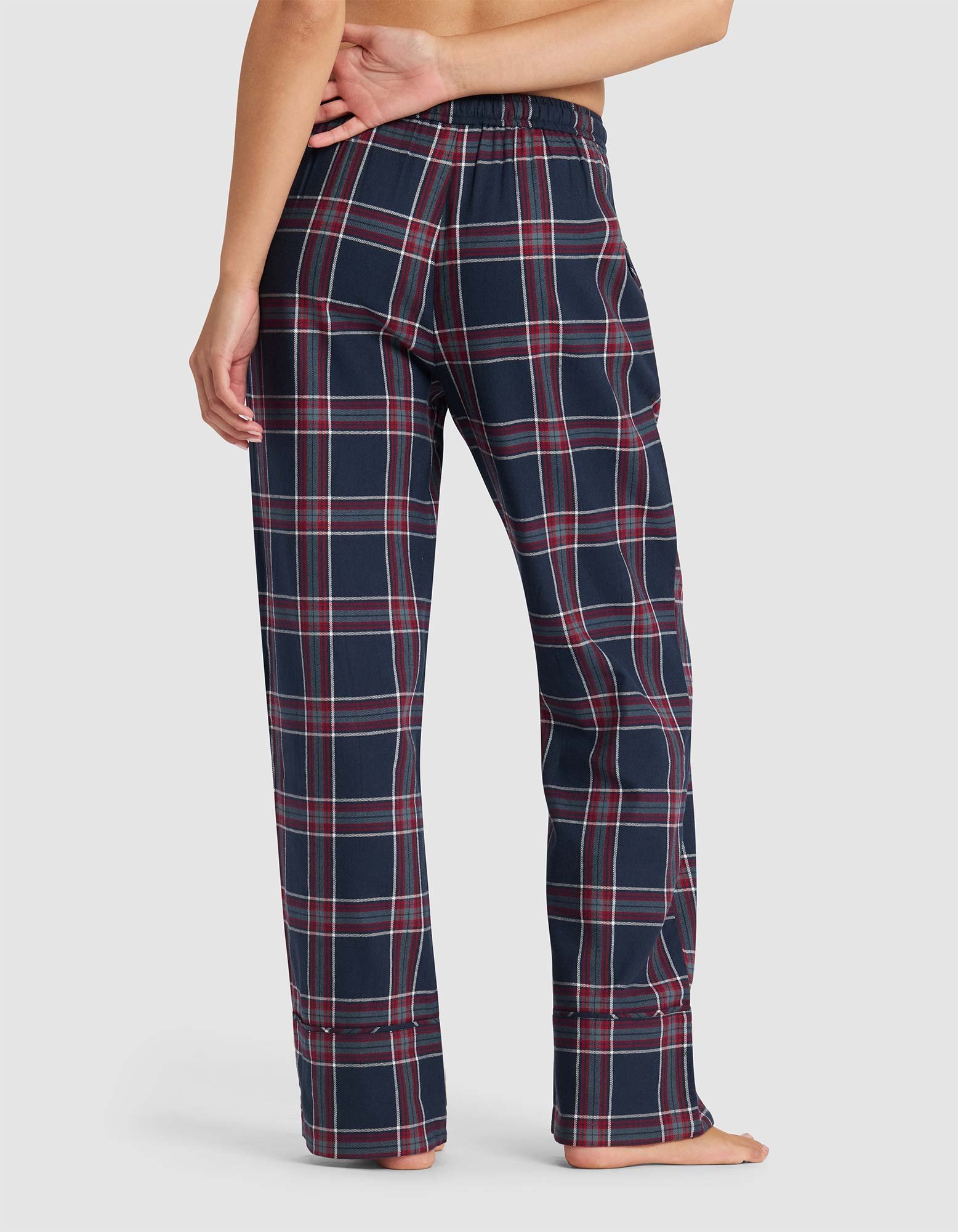 A woman stands barefoot, viewed from behind, wearing Cozy Earth Women's Farmhouse Pajama Pant in a loose-fitting navy blue plaid with red and white stripes.|Color:Eclipse Plaid