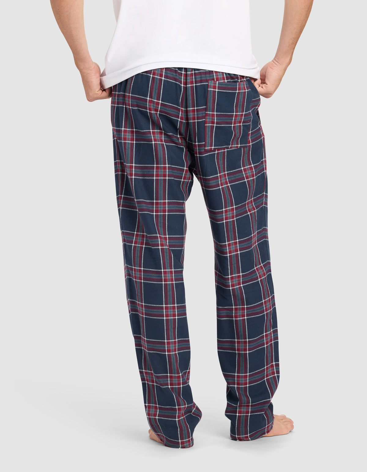 A person stands barefoot, facing away, wearing a white shirt and Cozy Earth Men’s Farmhouse Pajama Pant in navy blue with a red and white plaid pattern. The background is plain light gray.|Color:Eclipse Plaid
