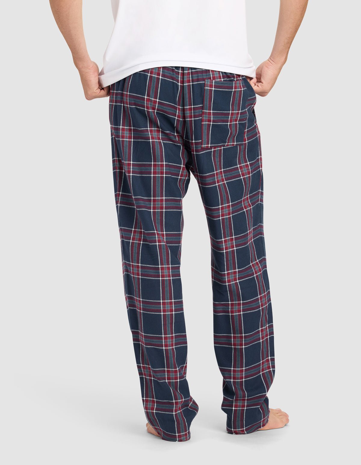A person stands barefoot, facing away, wearing a white shirt and Cozy Earth Men’s Farmhouse Pajama Pant in navy blue with a red and white plaid pattern. The background is plain light gray.