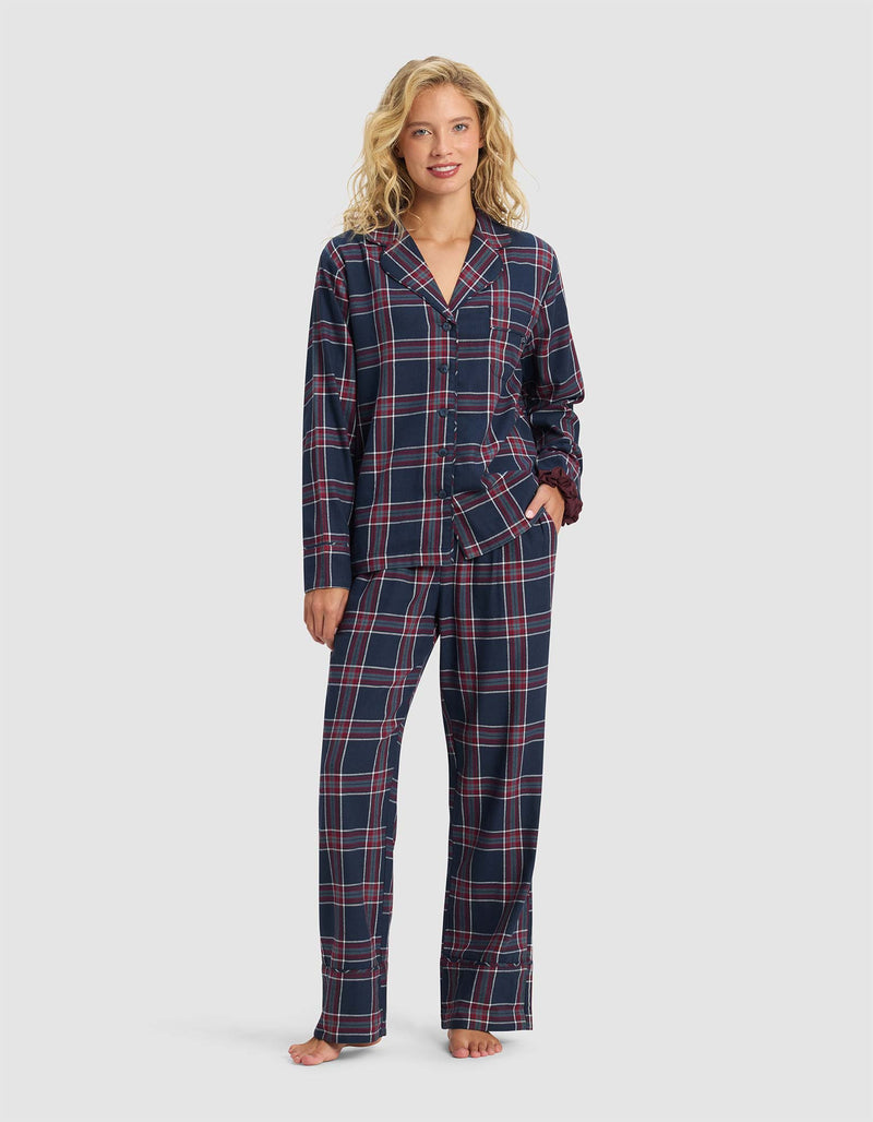 A woman stands barefoot, smiling in Cozy Earth's Women's Farmhouse Pajama Pant, featuring a navy blue base with red and white plaid. She pairs it with a matching long-sleeve button-up top, posing against a plain light background.
