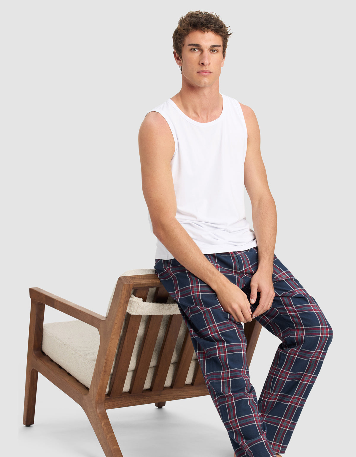 A young man in a white sleeveless shirt and Cozy Earth Men's Farmhouse Pajama Pant sits on the armrest of a wooden chair with a neutral expression against a light gray background.|Color:Eclipse Plaid