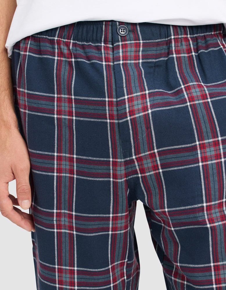 A person wears Cozy Earth Men's Farmhouse Pajama Pant, featuring blue, red, and white plaid, an elastic waistband, and a button. Their left hand rests beside their hip.
