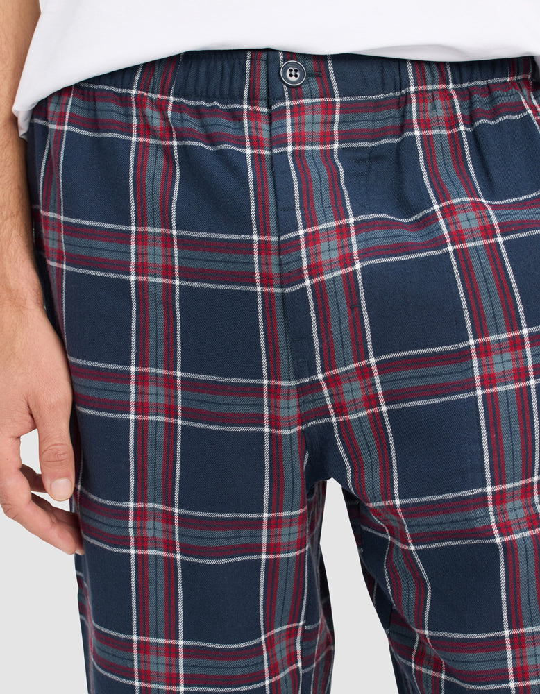 A person wears Cozy Earth Men's Farmhouse Pajama Pant, featuring blue, red, and white plaid, an elastic waistband, and a button. Their left hand rests beside their hip.|Color:Eclipse Plaid