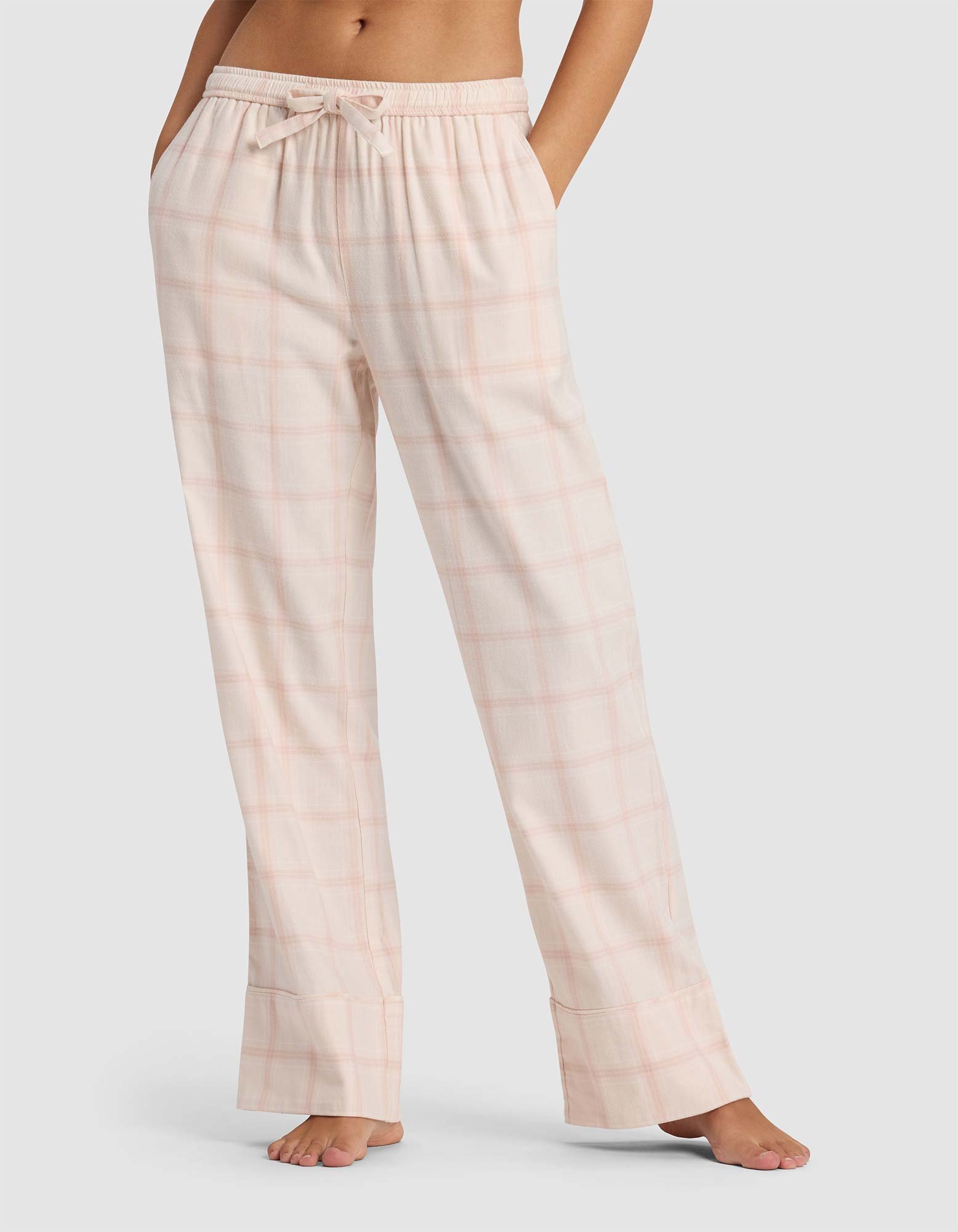 A woman models the Cozy Earth Women's Farmhouse Pajama Pant in light pink plaid, featuring a drawstring waist, while standing barefoot against a plain light background.|Color:Quartz Plaid
