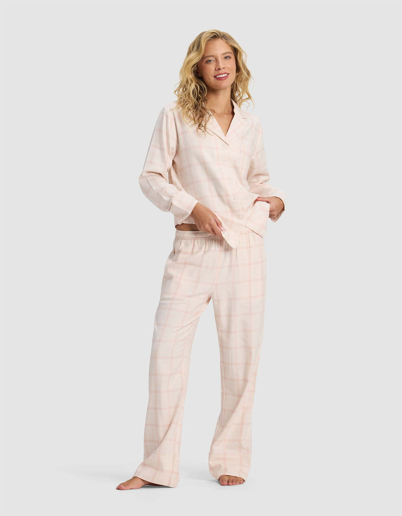 A woman with blonde hair wears Cozy Earth's Women's Farmhouse Pajama Pant in light pink plaid, standing barefoot and smiling against a plain light gray background.