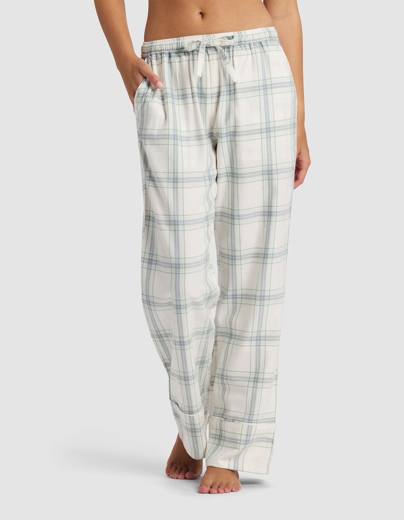 A woman wears Cozy Earth's Women's Farmhouse Pajama Pant—loose-fitting, light plaid with a drawstring waist and pockets—standing barefoot against a plain background.|Color:Wintergreen Plaid