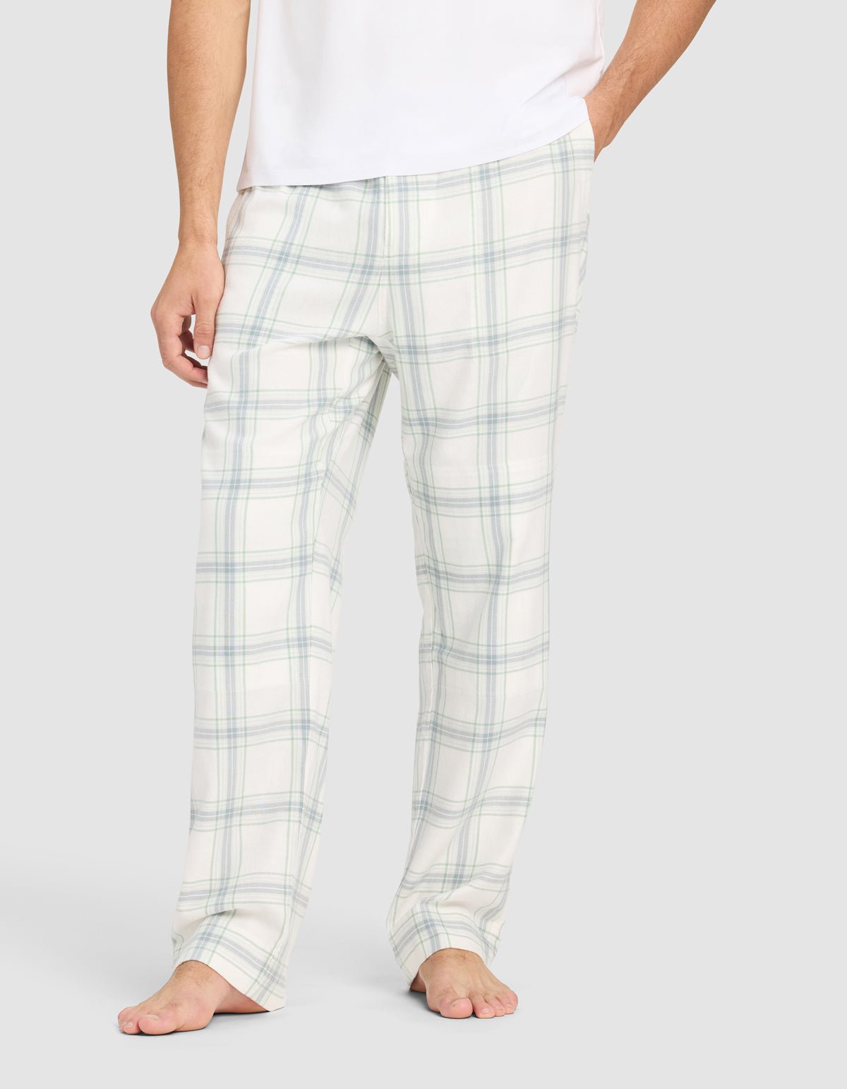 A person models the Cozy Earth Men's Farmhouse Pajama Pant—light-colored with blue and green plaid—standing barefoot, one hand in their pocket, against a plain background.