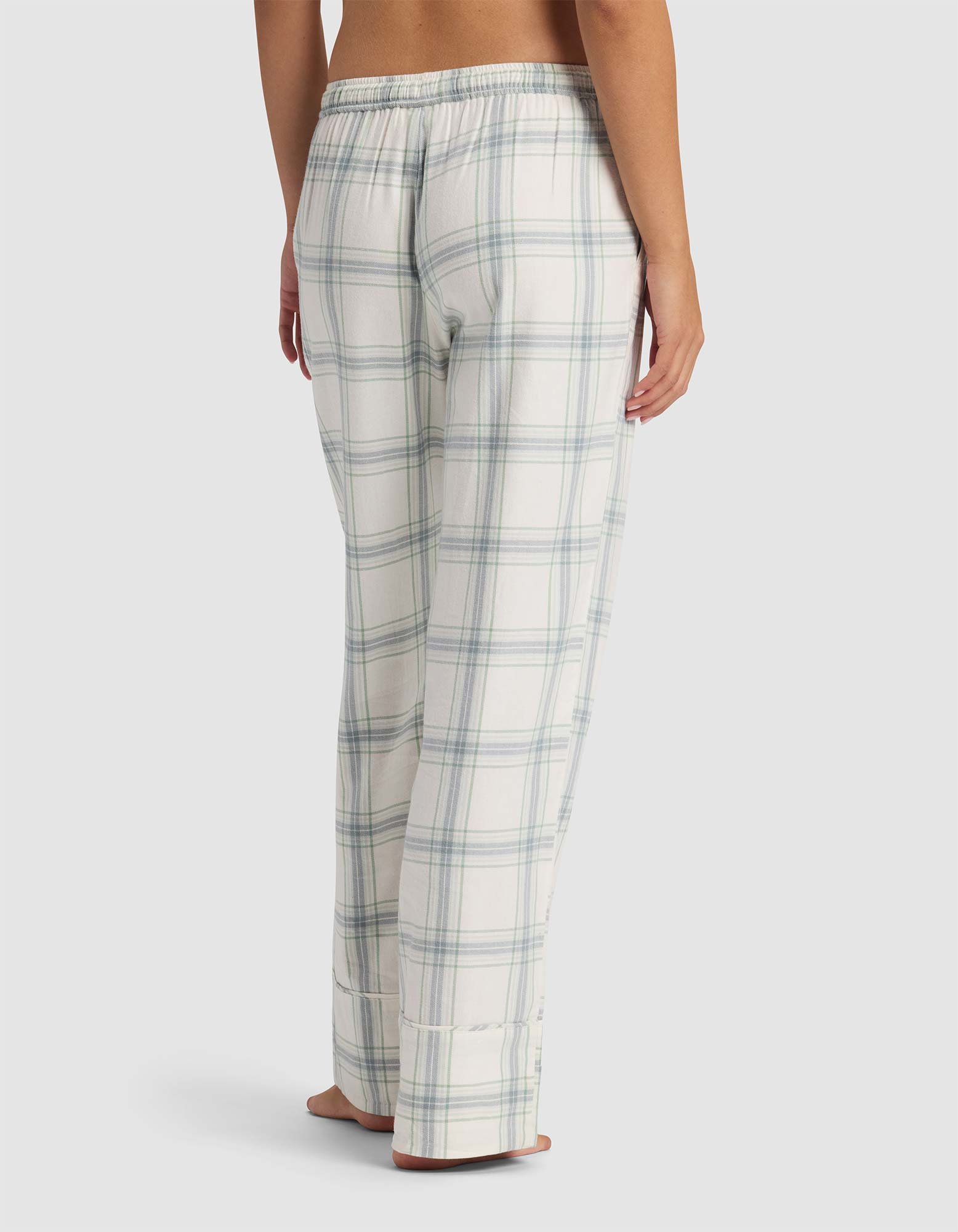 A woman stands barefoot, facing away, wearing Cozy Earth Women's Farmhouse Pajama Pant in white with light blue and gray plaid, featuring a relaxed fit and an elastic waistband.