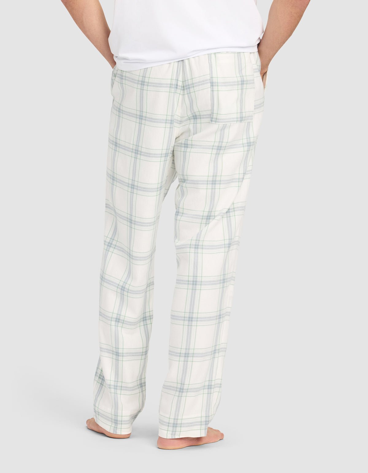 A person stands barefoot facing away in Cozy Earth Men's Farmhouse Pajama Pant—white with a light blue and green plaid pattern—and a white shirt, hands in pockets, against a plain light gray background.