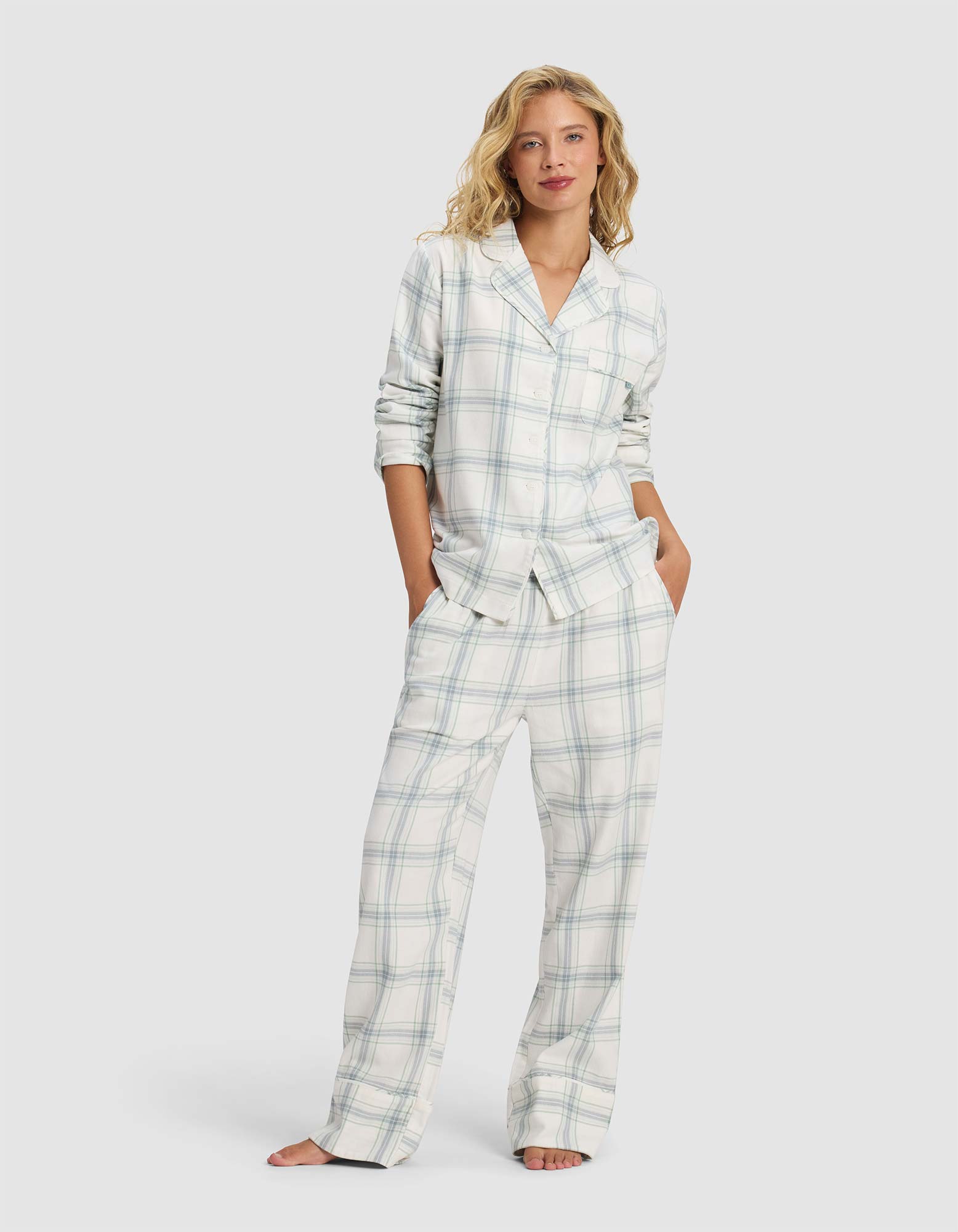 A woman with blonde wavy hair stands barefoot, hands in pockets, wearing the Cozy Earth Women's Farmhouse Pajama Pant—a white set with light blue and gray plaid—facing forward with a relaxed look.