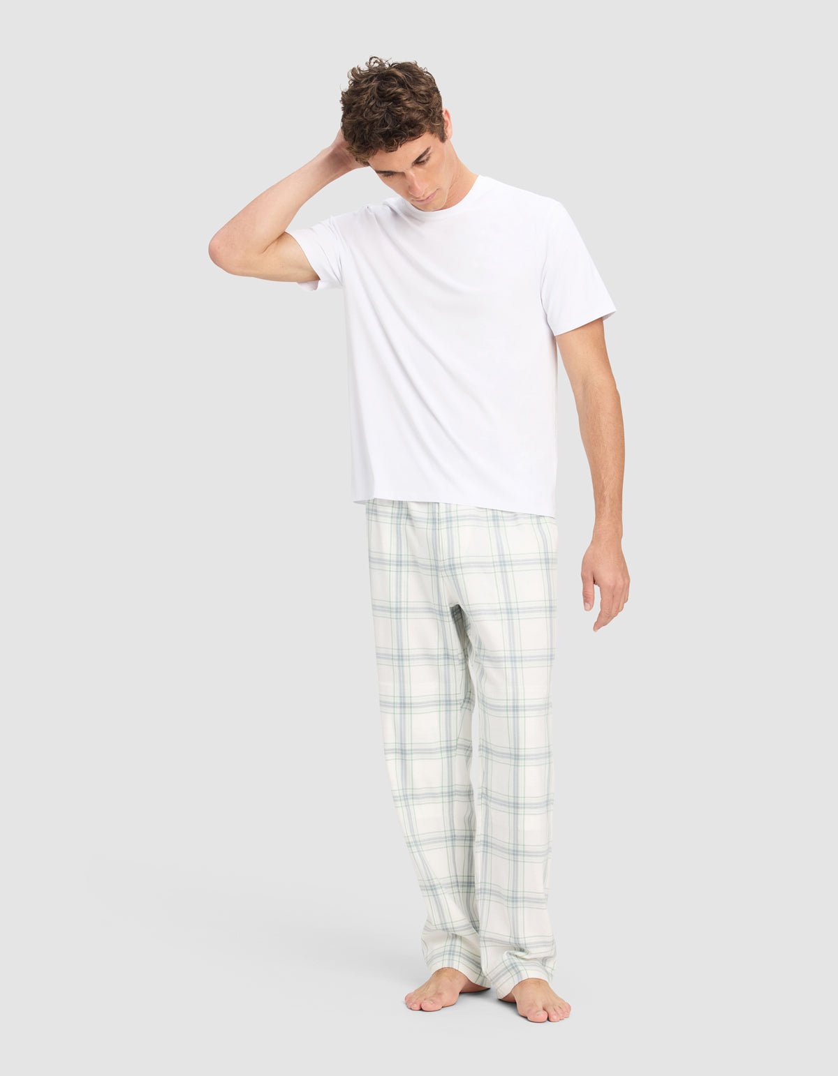A man stands barefoot on a plain background wearing Cozy Earth's Men's Farmhouse Pajama Pant and a white T-shirt, with one hand behind his head and looking downward.|Color:Wintergreen Plaid