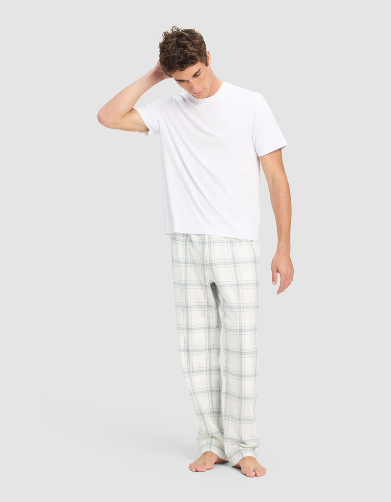A man stands barefoot on a plain background wearing Cozy Earth's Men's Farmhouse Pajama Pant and a white T-shirt, with one hand behind his head and looking downward.