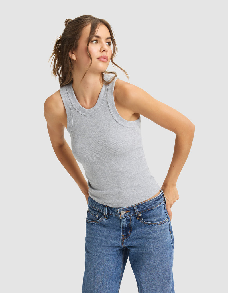 A woman with brown hair in a loose ponytail wears Cozy Earth's Women's Fine Ribbed Tank in gray and blue jeans, standing against a light background and looking slightly left with her hands on her hips.