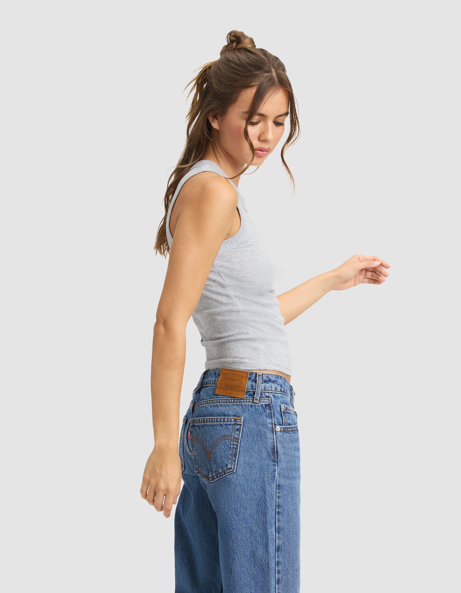 A young woman with long brown hair, styled half up, wears the Cozy Earth Women's Fine Ribbed Tank in light gray and high-waisted blue jeans. She stands sideways against a plain background, eyes closed and one arm slightly raised.|Color:Heather Grey