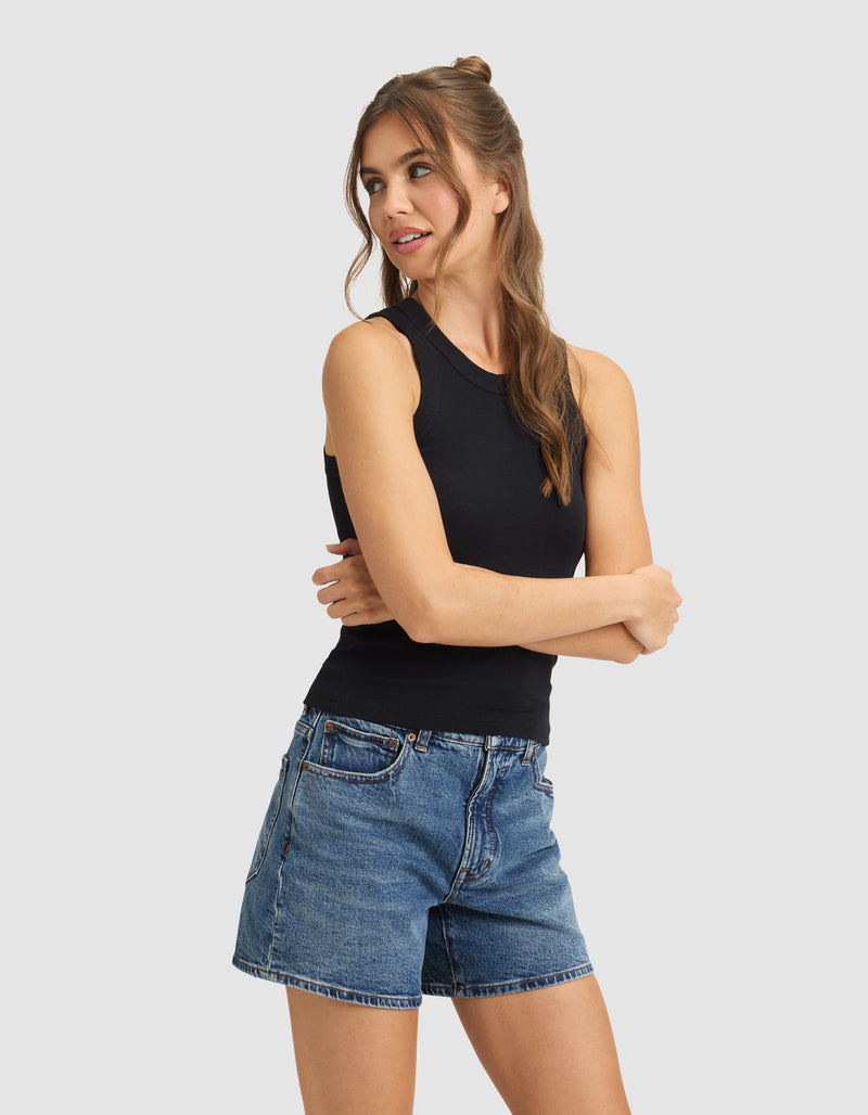 A woman with long brown hair models the Cozy Earth Women's Fine Ribbed Tank in black, paired with blue denim shorts. She stands with arms crossed, looking left against a plain light gray background.