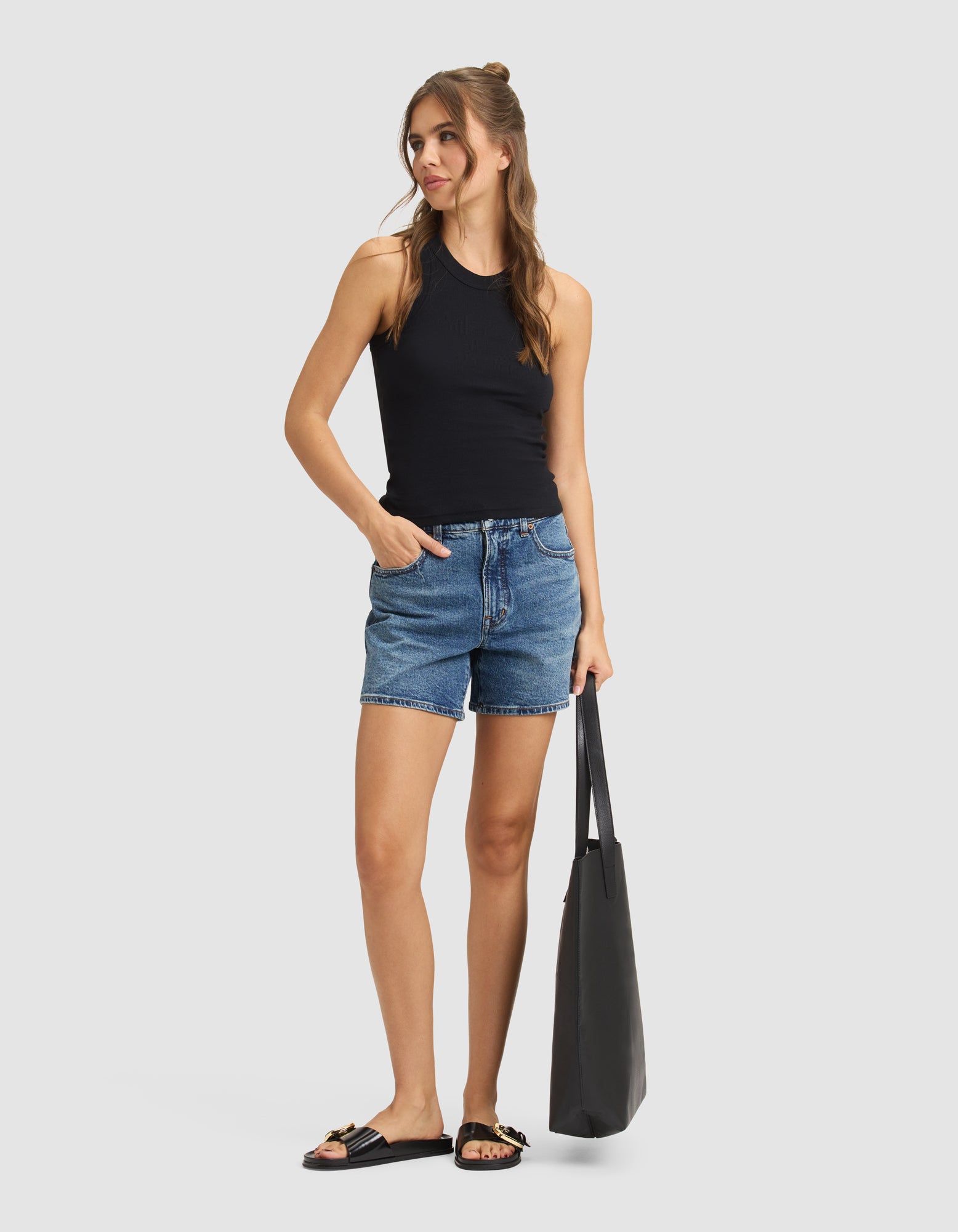 A woman stands against a plain background wearing the Cozy Earth Women's Fine Ribbed Tank in black, paired with denim shorts, black sandals with gold accents, and holding a large black tote bag. She has long hair and looks to the side with one hand in her pocket.
