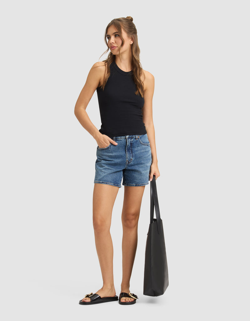 A woman stands against a plain background wearing the Cozy Earth Women's Fine Ribbed Tank in black, paired with denim shorts, black sandals with gold accents, and holding a large black tote bag. She has long hair and looks to the side with one hand in her pocket.