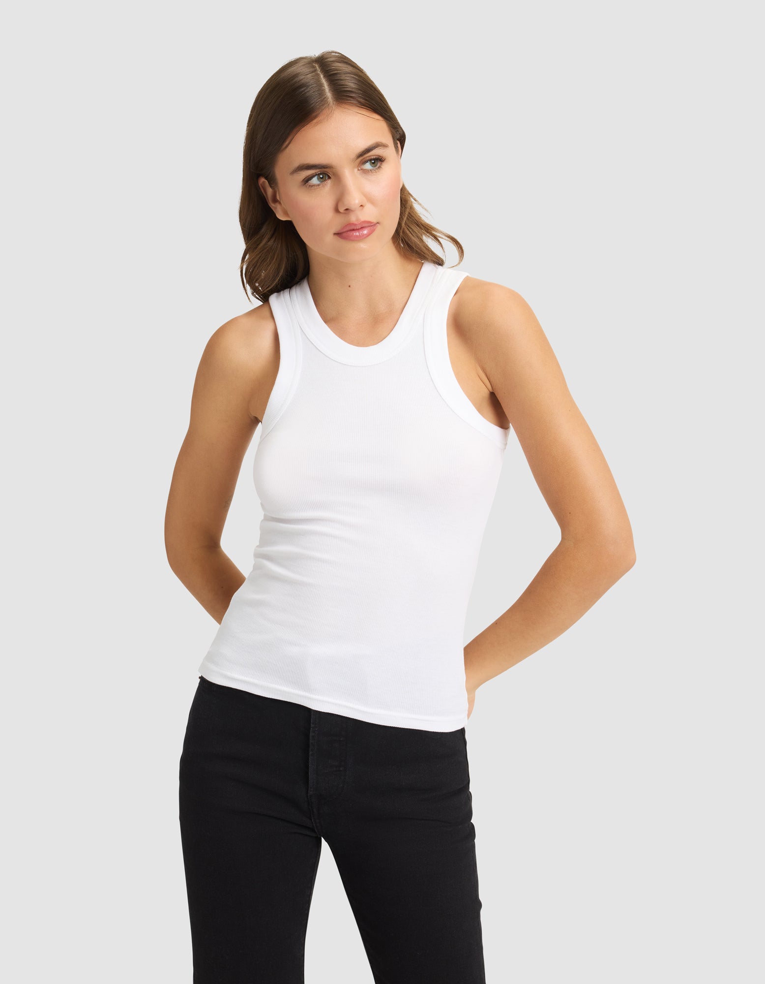 A woman with long brown hair models the Cozy Earth Women's Fine Ribbed Tank in white, paired with black pants. She stands against a plain light gray background, looking to her right with her hands behind her back.
