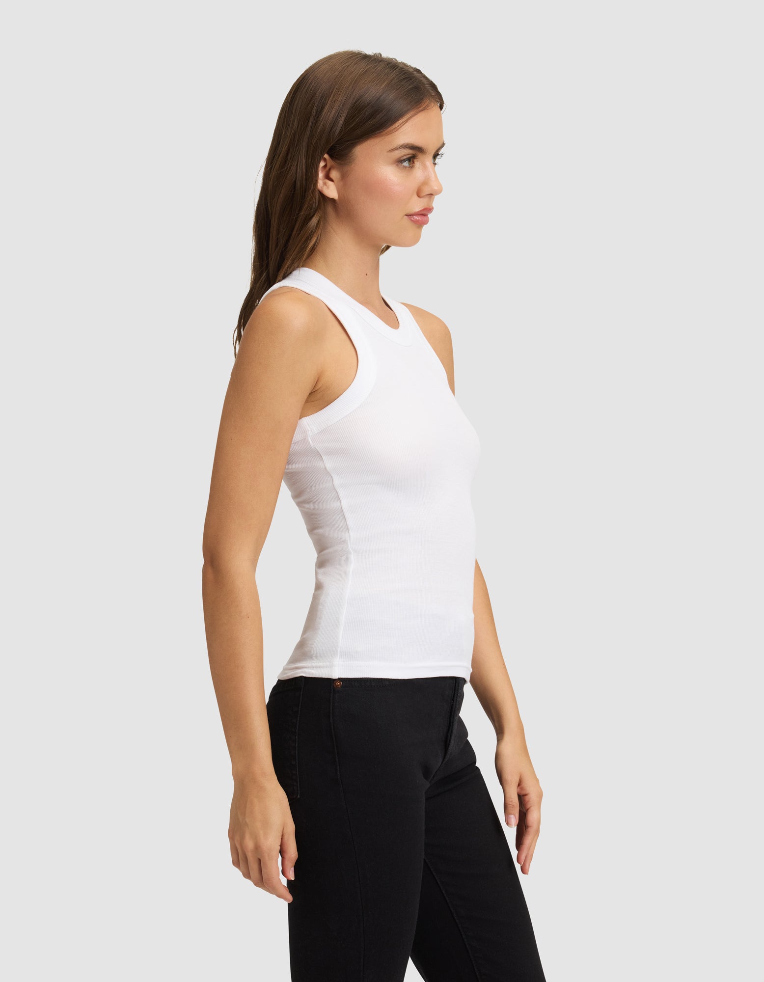 A woman with long brown hair stands in profile, wearing Cozy Earth's Women's Fine Ribbed Tank in white and black pants against a plain light gray background.
