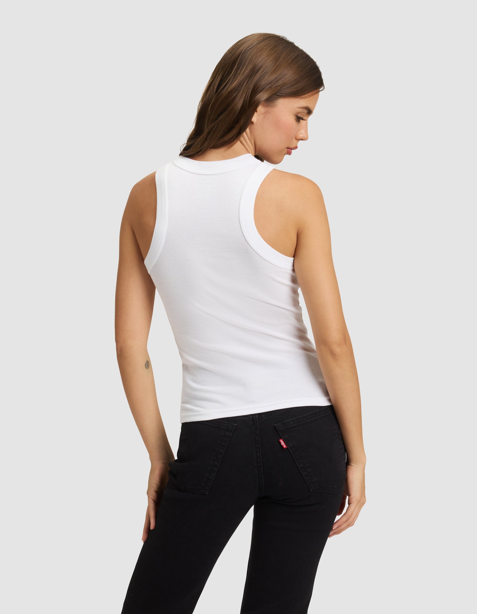 A woman with long brown hair is shown from the back wearing the Cozy Earth Women's Fine Ribbed Tank and black jeans, standing against a plain light gray background.