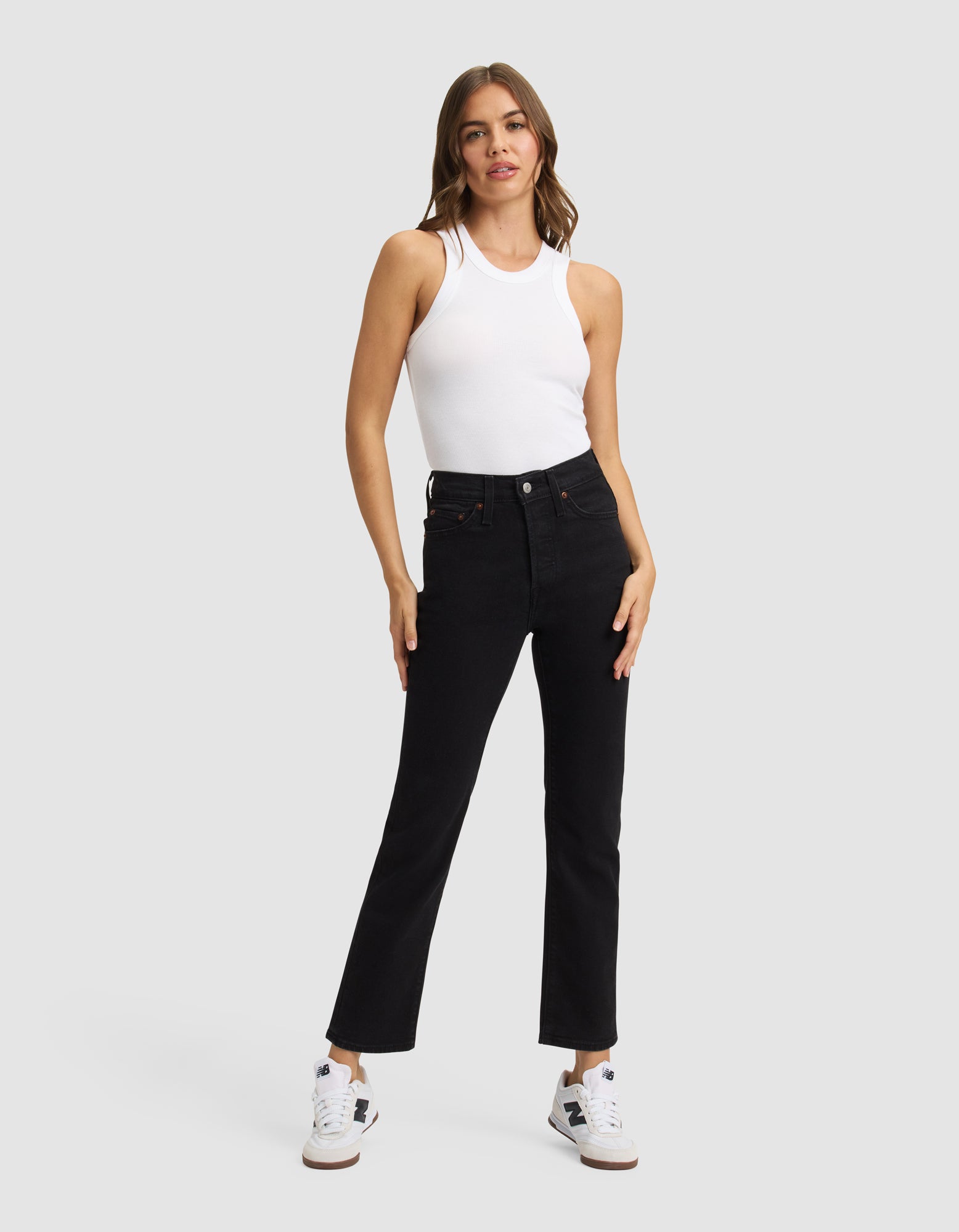 A woman with long brown hair poses against a plain background wearing the Cozy Earth Women's Fine Ribbed Tank in white, paired with black straight-leg jeans and white sneakers. She stands with one hand on her hip, looking at the camera.