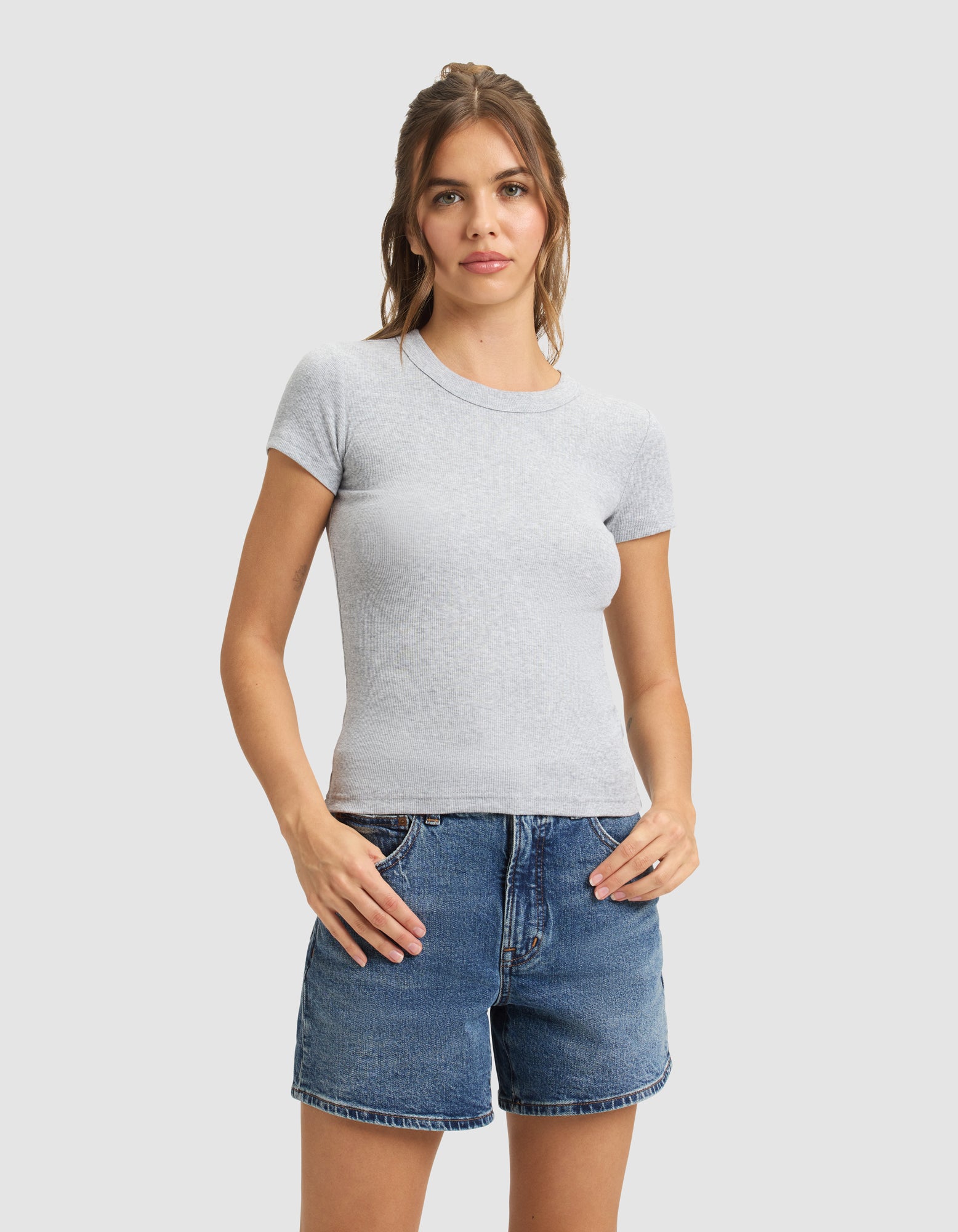 A woman stands against a light gray background wearing the Cozy Earth Women's Fine Ribbed Tee in light gray and blue denim shorts, with her hands on her hips. 