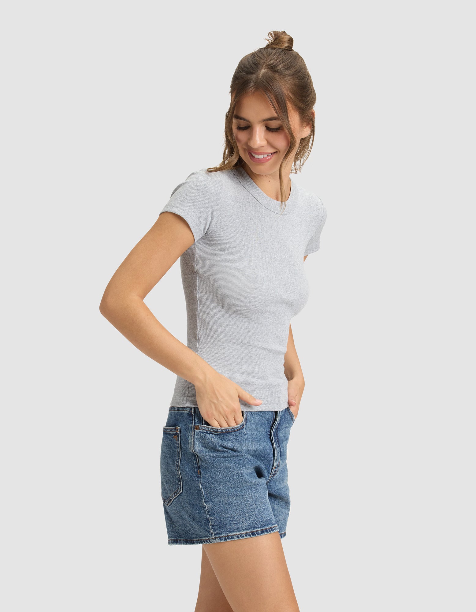 A woman with brown hair smiles with her hands in her pockets, wearing the Cozy Earth Women's Fine Ribbed Tee in light gray and blue denim shorts, standing against a plain light background. 