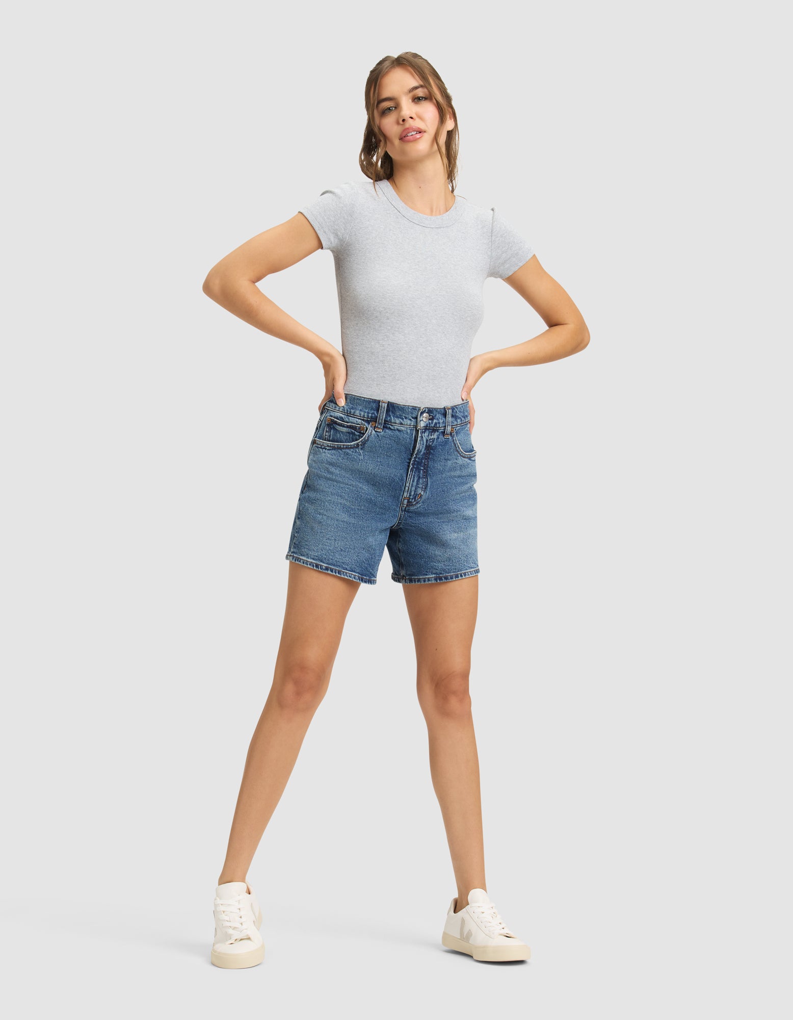 A woman stands confidently with hands on hips, wearing the Cozy Earth Women's Fine Ribbed Tee in light gray, paired with blue denim shorts and white sneakers against a plain light gray background. 