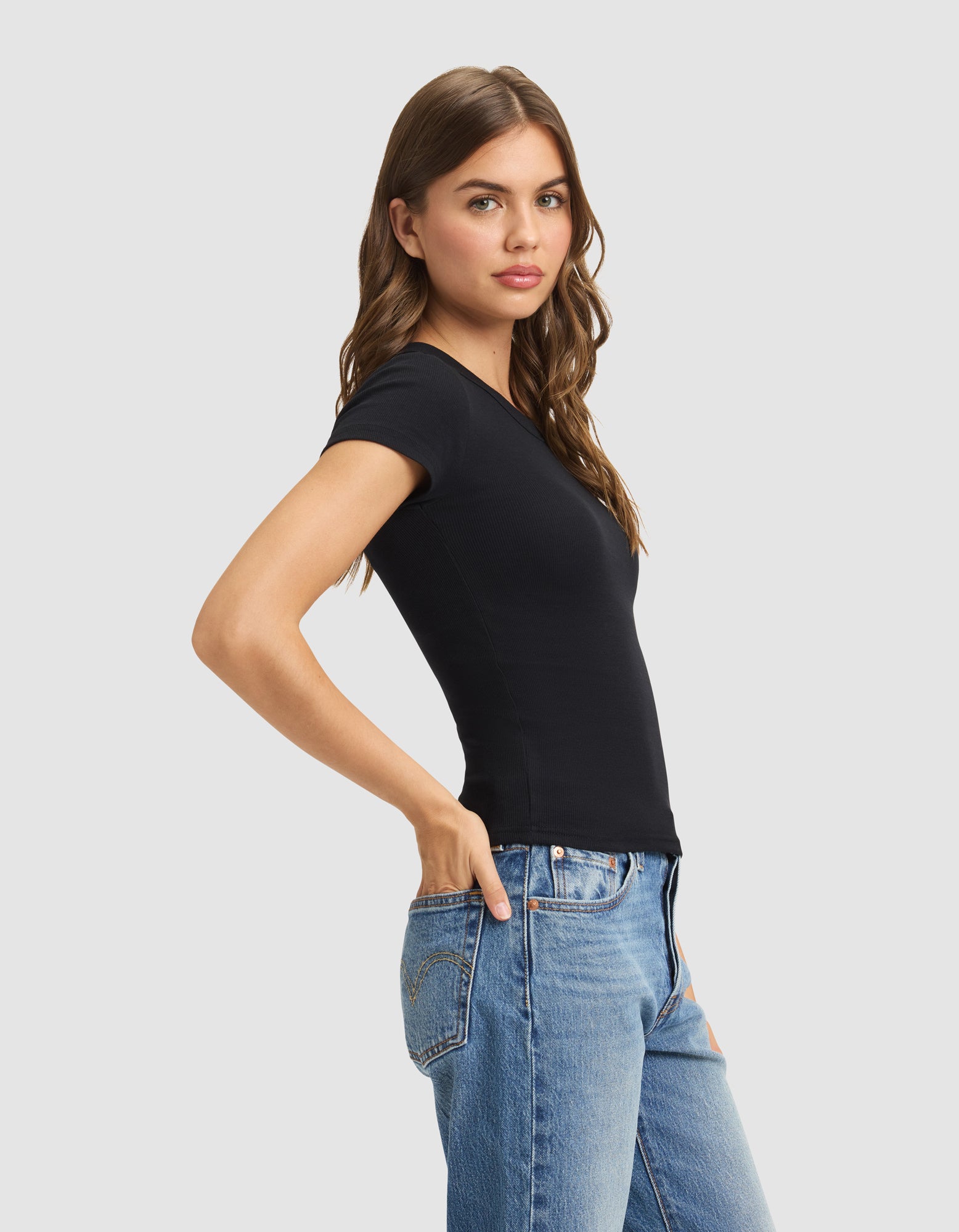 A woman with long brown hair wears the Women's Fine Ribbed Tee by Cozy Earth in black and blue jeans, standing sideways with one hand in her pocket against a plain light background. 