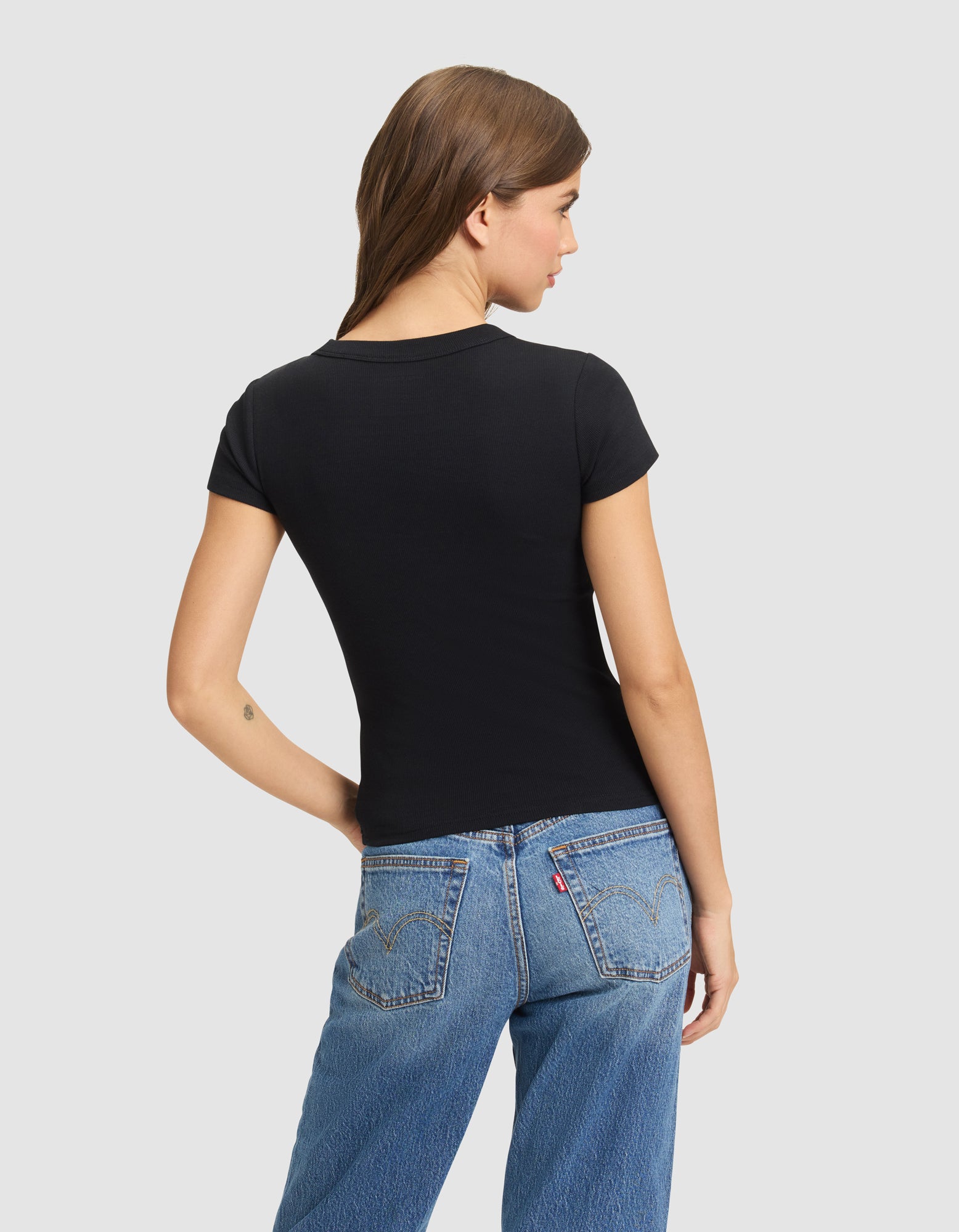 A woman with brown hair wears the Cozy Earth Women's Fine Ribbed Tee in black and blue jeans, standing facing away against a plain light gray background. 