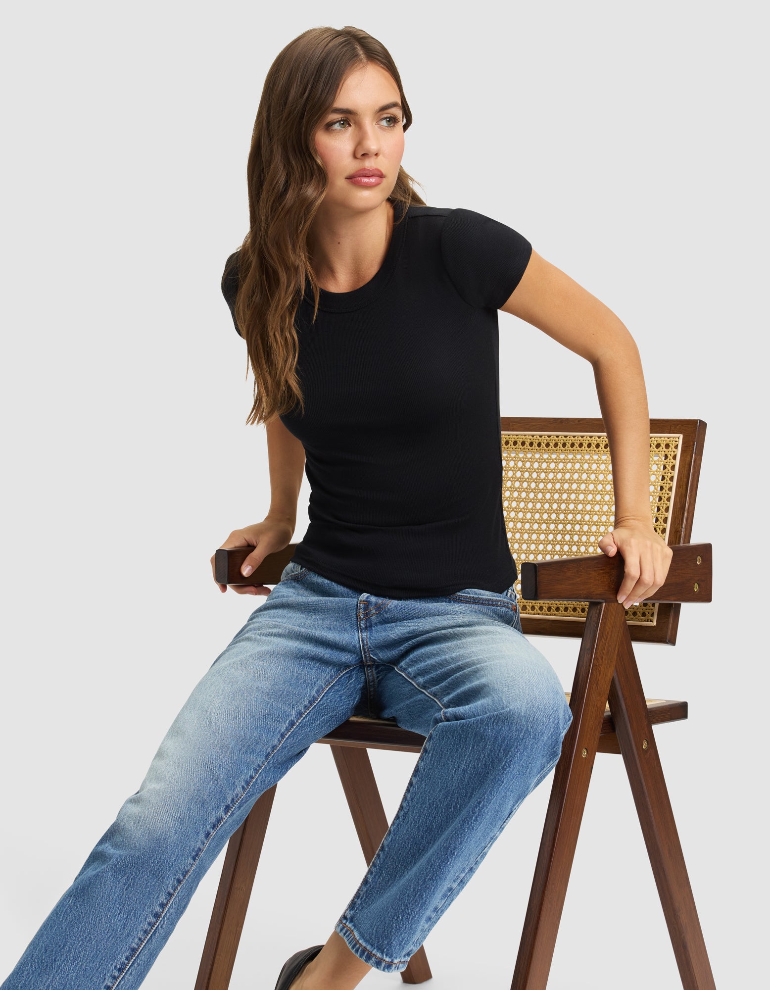 A woman with long brown hair, wearing the Cozy Earth Women's Fine Ribbed Tee and blue jeans, sits sideways on a wooden chair with a woven backrest, looking to the side against a plain light background. 