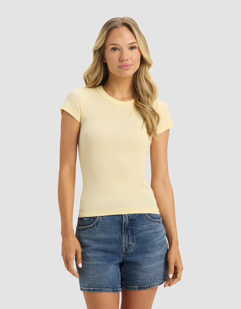 A woman with long blonde hair is wearing the Cozy Earth Women's Fine Ribbed Tee in light yellow and denim shorts, standing against a plain light grey background.