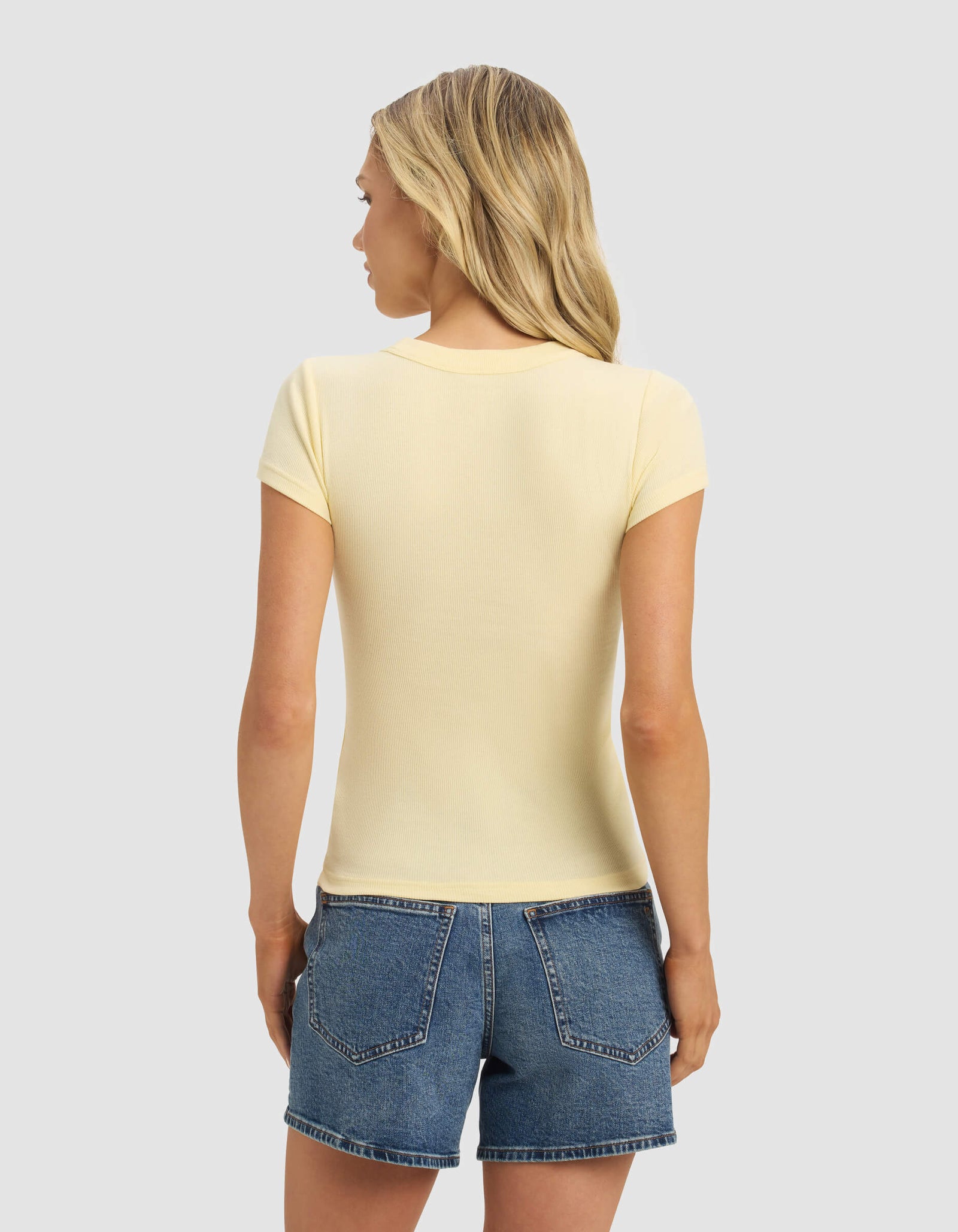 A woman with long blonde hair is seen from behind wearing the Cozy Earth Women's Fine Ribbed Tee in light yellow and blue denim shorts, set against a plain light gray background.