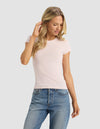 A woman with long blonde hair models the Cozy Earth Women's Fine Ribbed Tee in light pink, paired with blue jeans, as she stands against a plain light gray background, looking down and gently touching her hair.|Color:Ballet