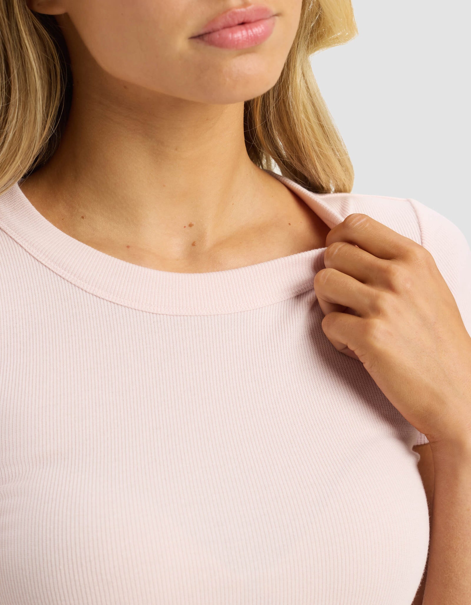 A woman wears the Cozy Earth Women's Fine Ribbed Tee in light pink, gently pulling at the collar with her right hand. Only her lower face, neck, and upper torso are visible against a plain light background.