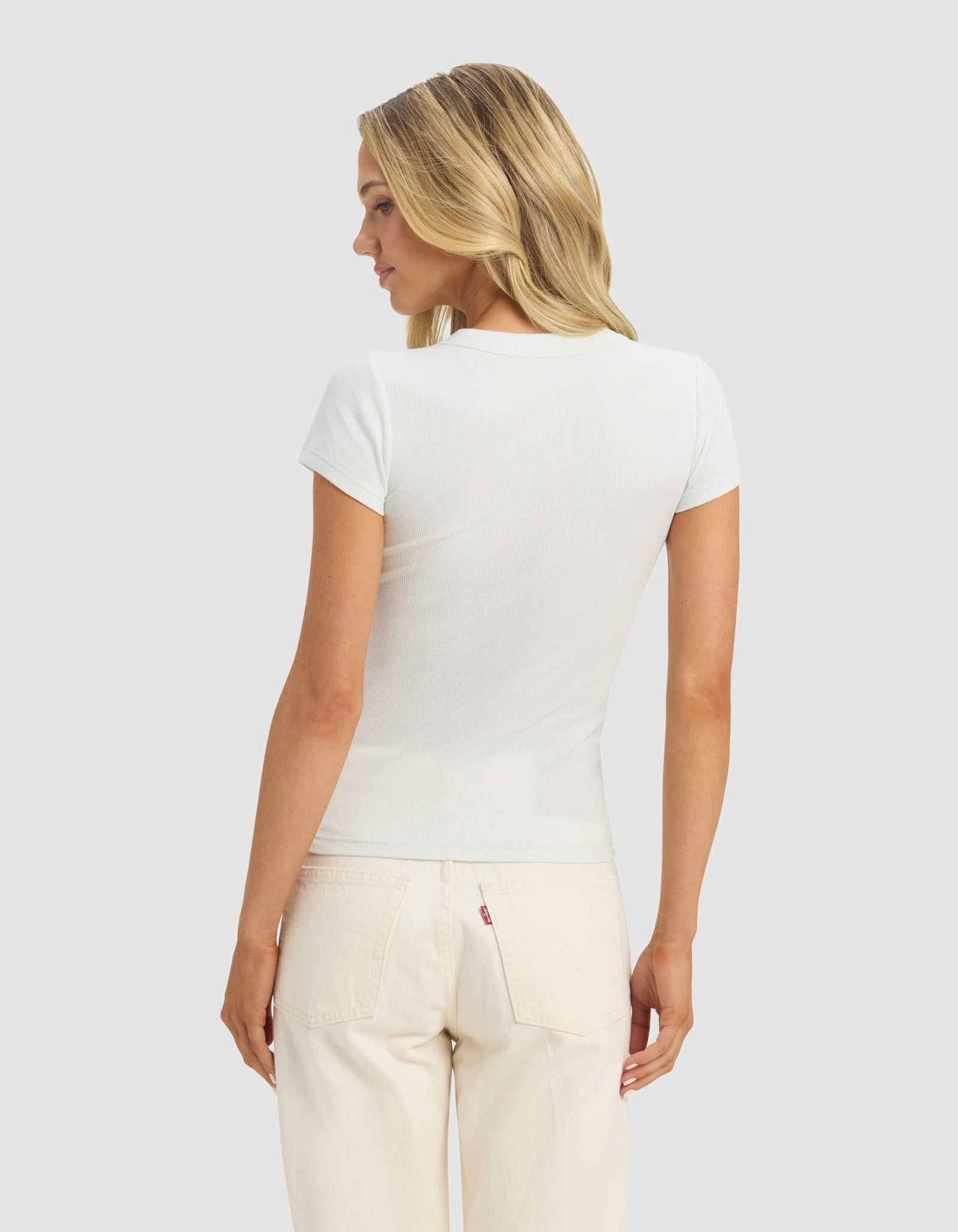 A woman with blonde hair faces away, wearing the Cozy Earth Women's Fine Ribbed Tee in white and light-colored jeans, set against a plain light gray background.