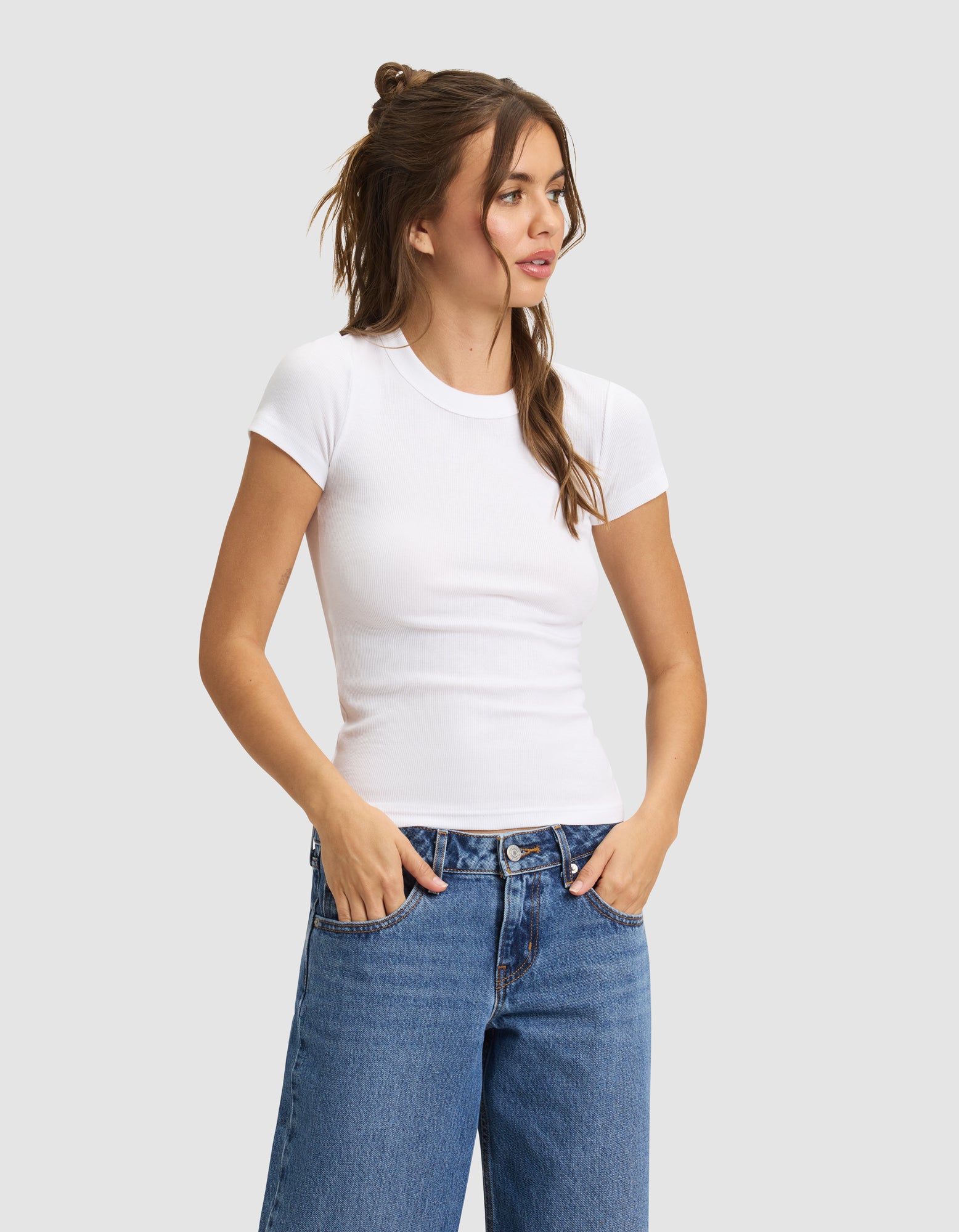 A woman with long brown hair in a half-up style wears the Cozy Earth Women's Fine Ribbed Tee in white and blue jeans. She stands with hands in pockets, looking to the side against a plain gray background. 