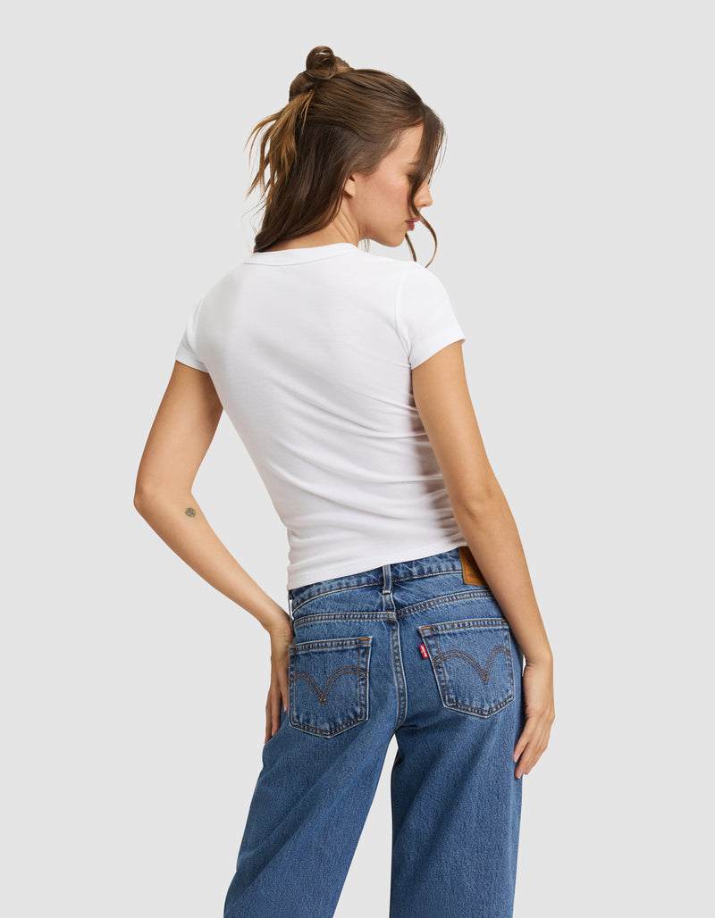 A woman with brown hair in a half-up style models the Cozy Earth Women's Fine Ribbed Tee, a fitted white short-sleeve shirt, paired with blue jeans as she faces away from the camera against a light gray background.