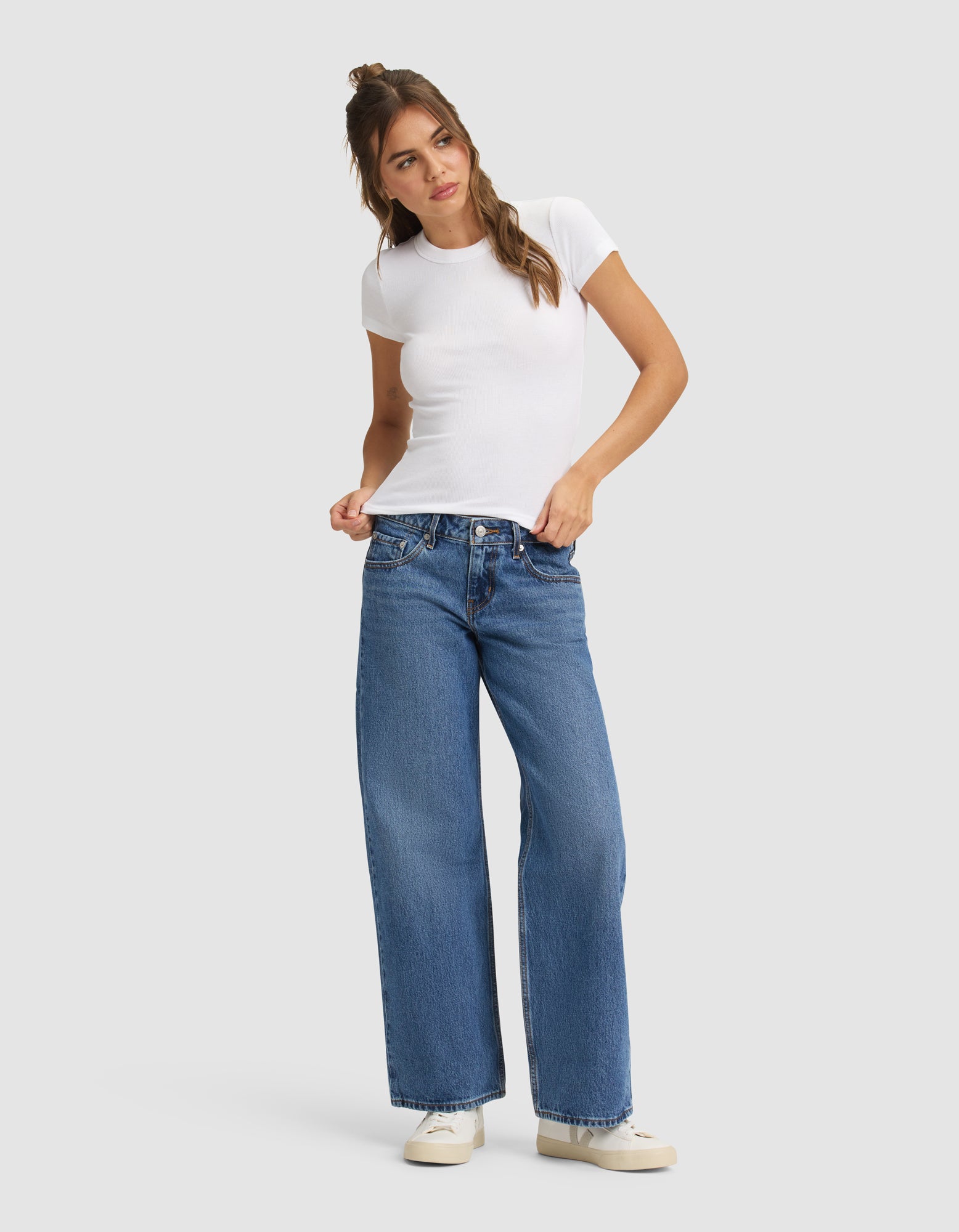 A woman with long brown hair styled half-up poses against a plain background, wearing the Cozy Earth Women's Fine Ribbed Tee, blue wide-leg jeans, and white sneakers. She stands with one hand in her pocket and the other on her waistband. 