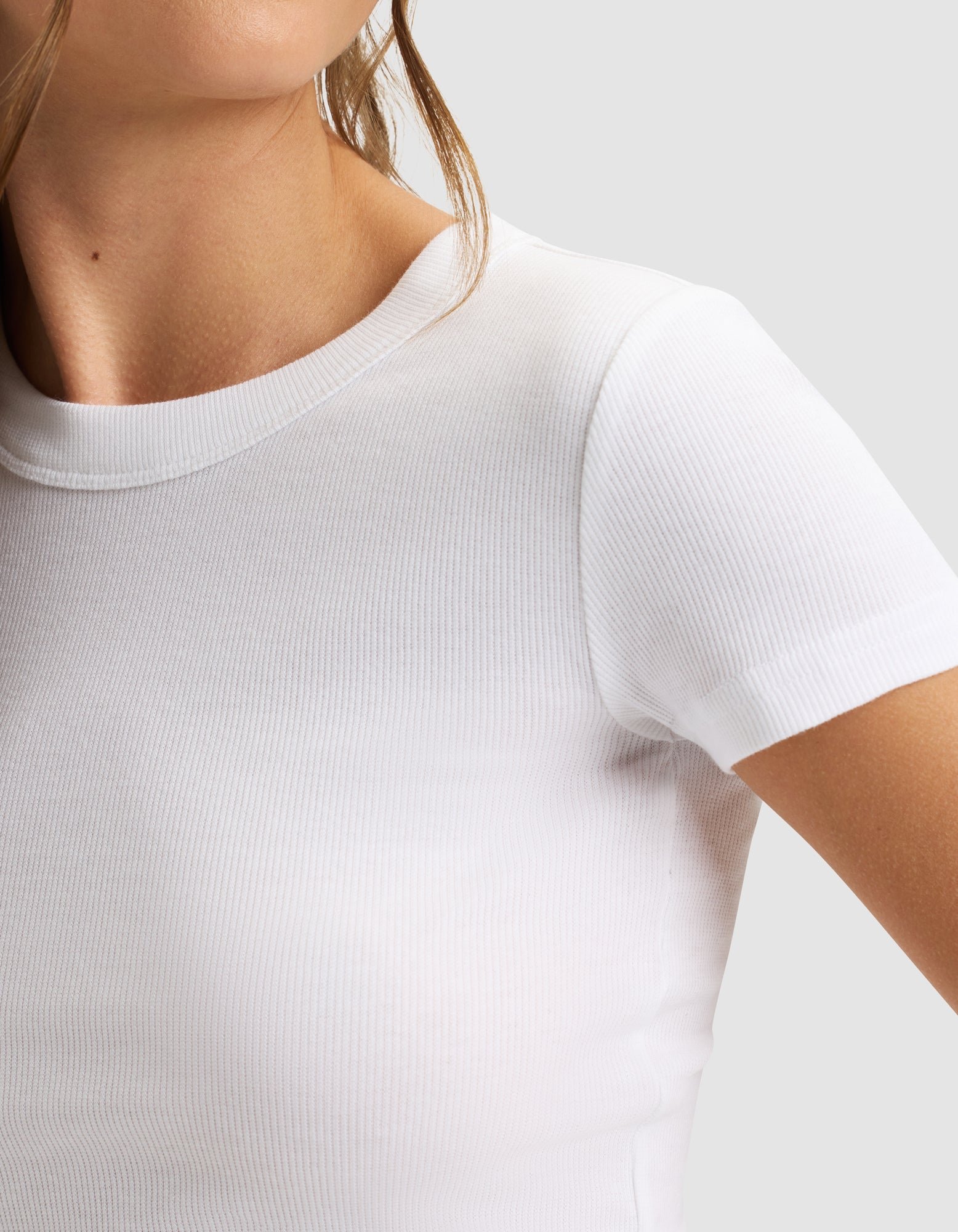 A close-up of a woman wearing the Cozy Earth Women's Fine Ribbed Tee in white, featuring fitted short sleeves and ribbed texture. The image highlights her upper chest, shoulder, and part of her face against a neutral background. 