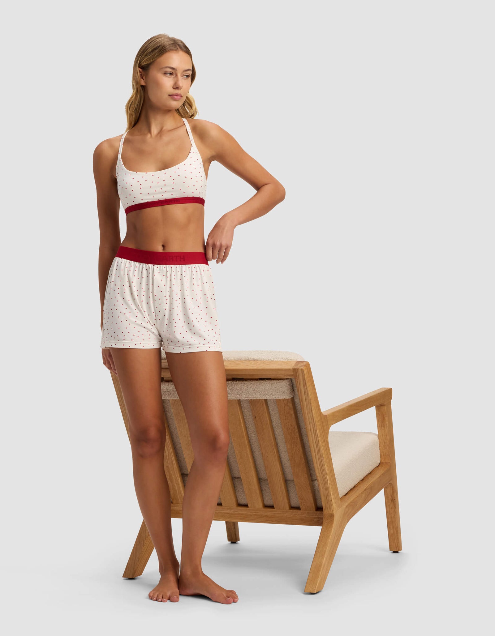 A woman in Cozy Earth's Women's First Layer Pajama Boxer Shorts stands barefoot beside a wooden armchair with light cushions, looking to her right.