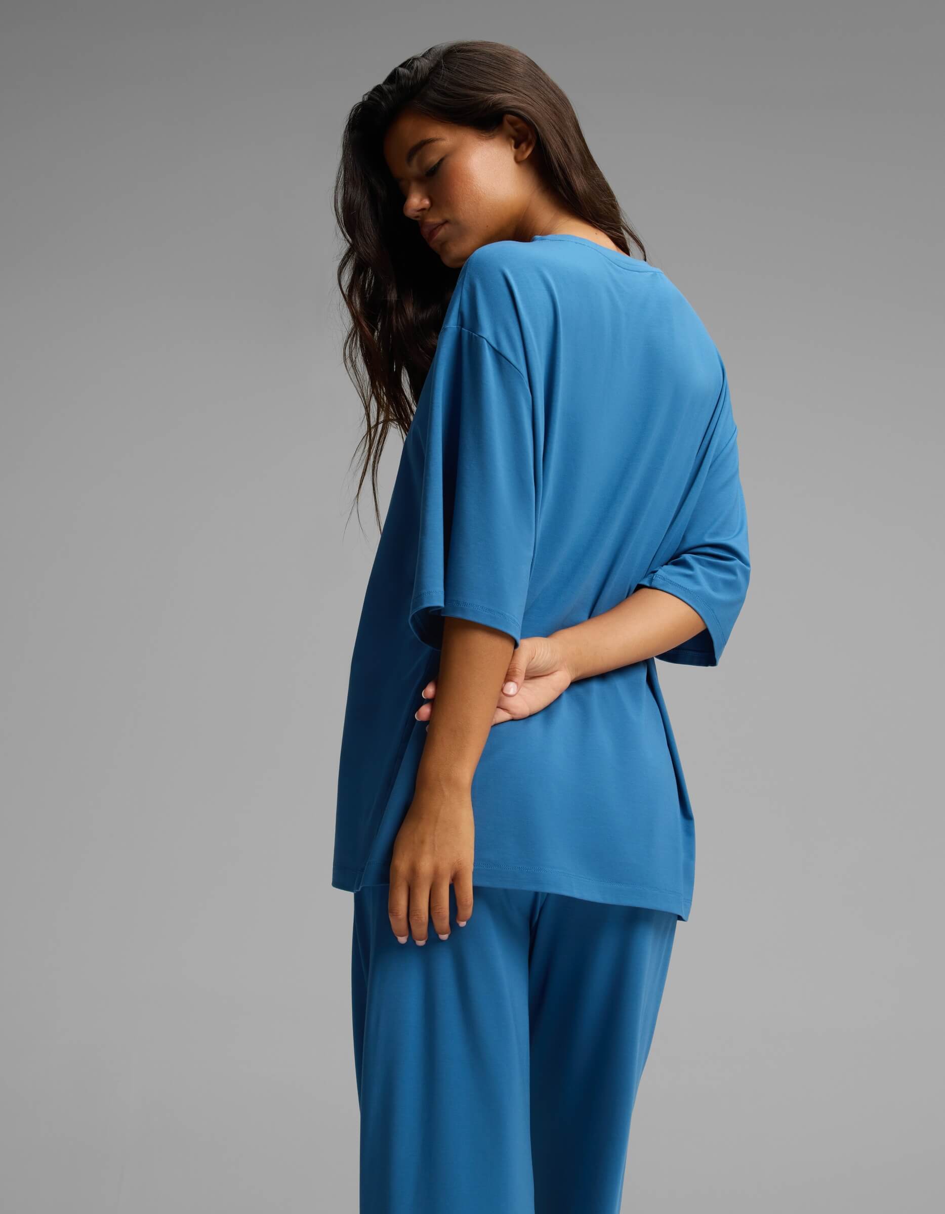 A person with long dark hair stands in profile against a plain gray background, wearing the Cozy Earth Women’s First Layer Boyfriend Sleep Tee in blue, paired with matching pants. One arm is bent behind their back.|Color:Anchor