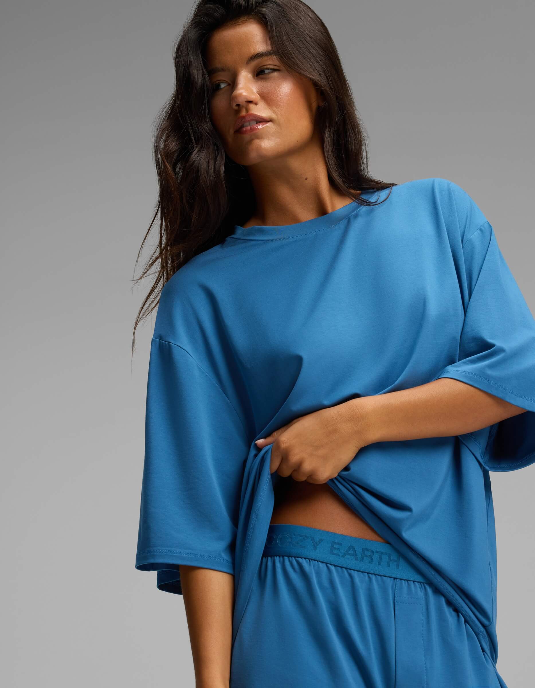 A woman with long dark hair gazes to the side against a plain gray background while lifting the hem of her loose blue Cozy Earth Women’s First Layer Boyfriend Sleep Tee to reveal matching blue Boxer Shorts featuring the “COZY EARTH” logo on the waistband.