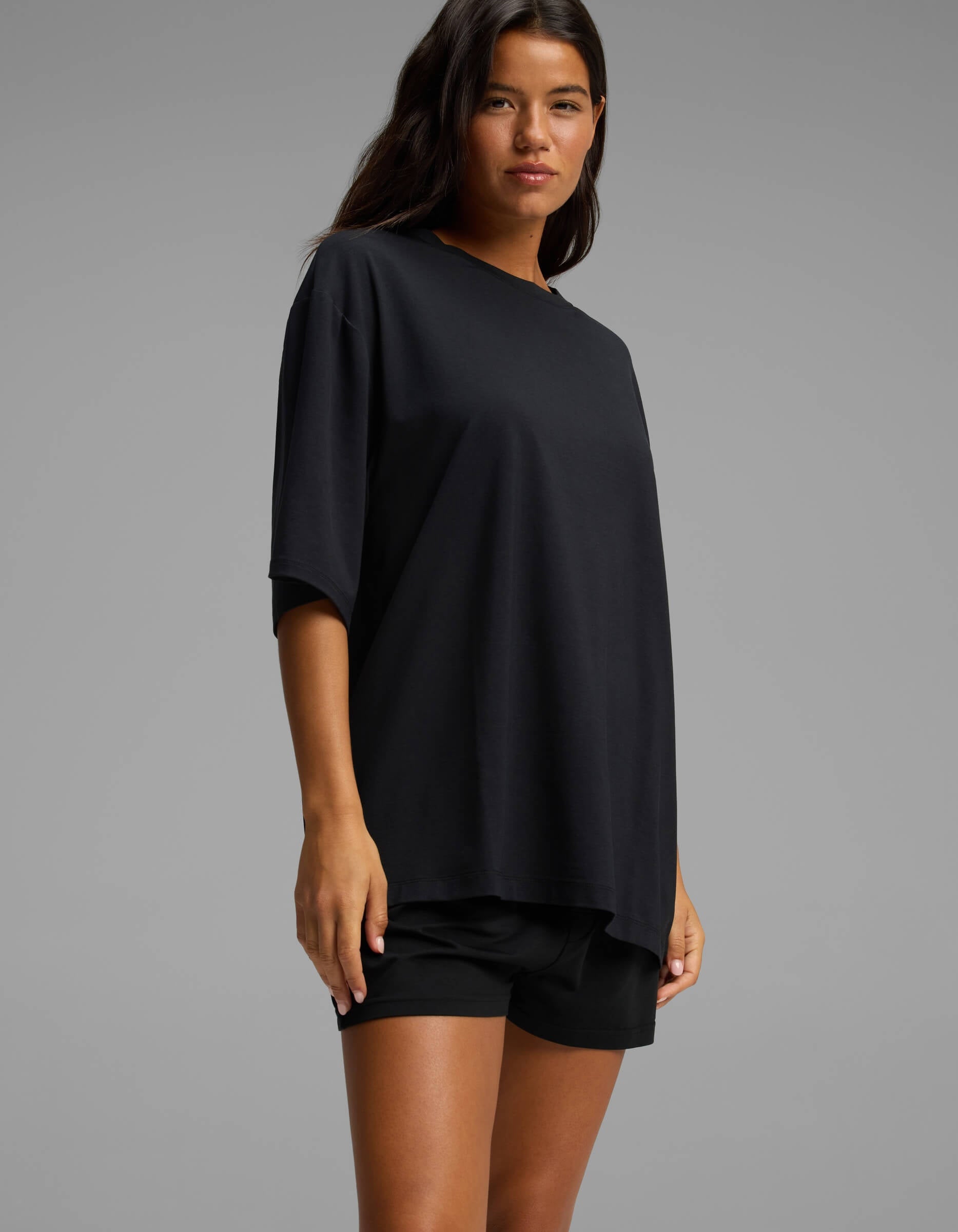A woman stands against a gray background, wearing the Cozy Earth Women’s First Layer Boyfriend Sleep Tee & Boxer Short Set in black, looking confidently at the camera with her hair down.