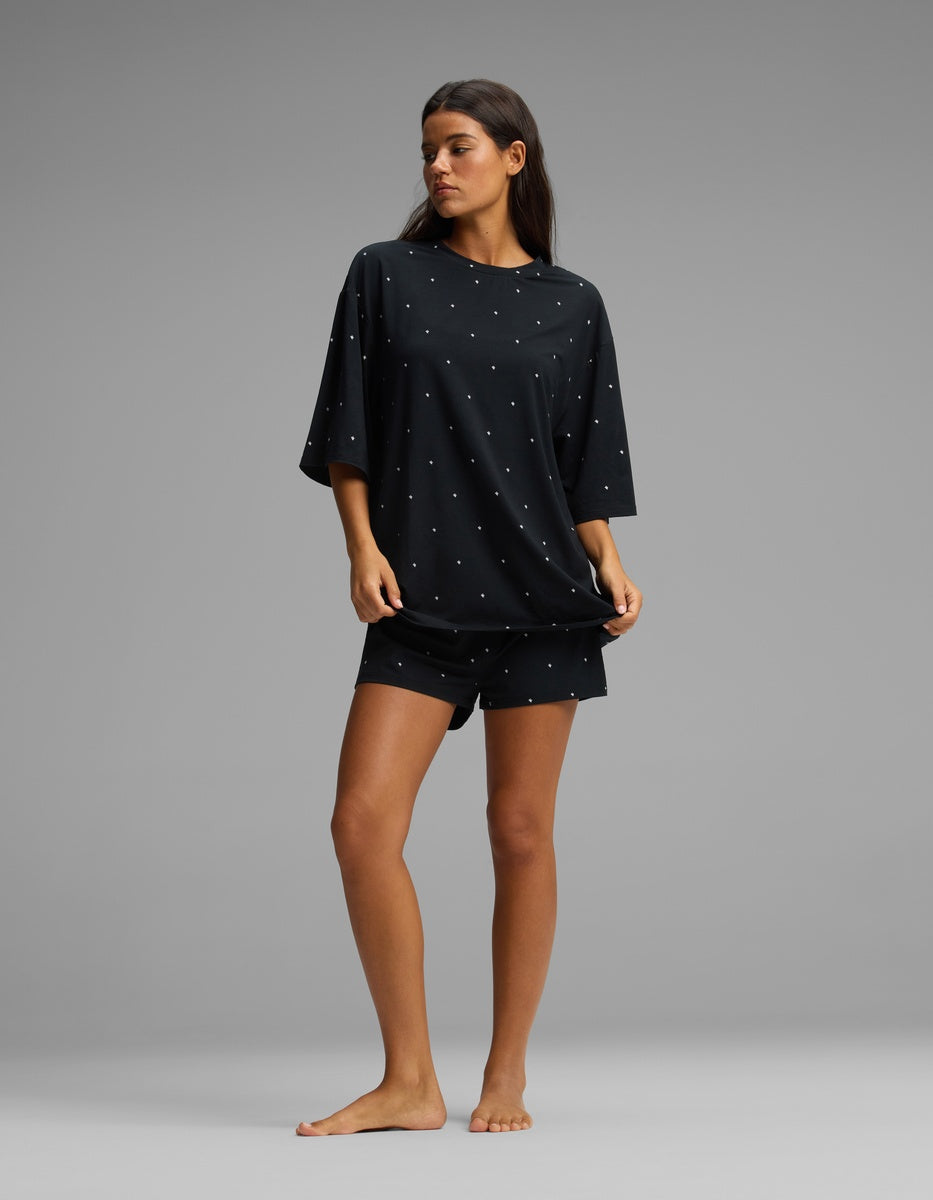 A woman stands barefoot against a gray background, wearing the Cozy Earth Women’s First Layer Boyfriend Sleep Tee & Boxer Short Set in a loose-fitting black fabric with small white dots.