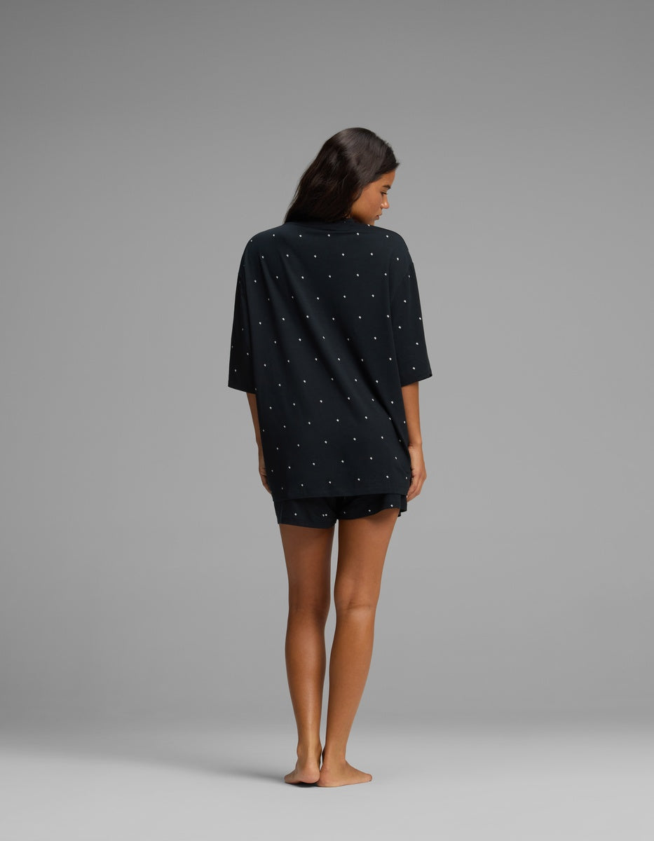 A woman stands barefoot on a gray background, facing away from the camera, wearing the Cozy Earth Women’s First Layer Boyfriend Sleep Tee & Boxer Short Set in a dark color with small white dots.