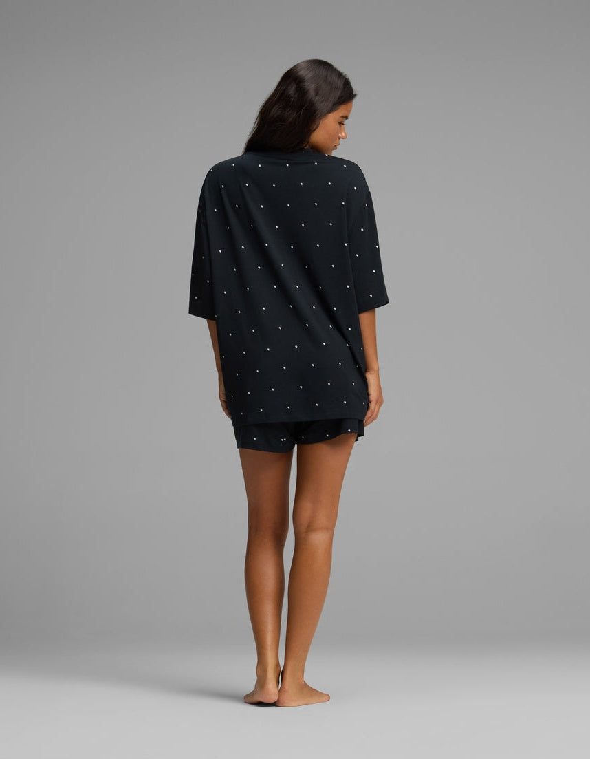 A woman stands barefoot on a gray background, facing away from the camera, wearing the Cozy Earth Women’s First Layer Boyfriend Sleep Tee & Boxer Short Set in a dark color with small white dots.|Color:Mini Ghost