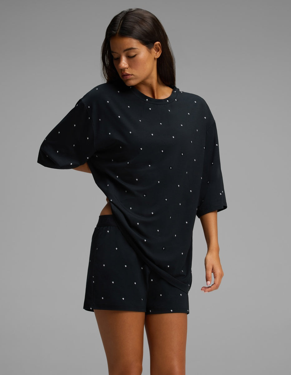 A woman stands against a gray background, looking down and gently holding the side of her shirt while wearing the Cozy Earth Women’s First Layer Boyfriend Sleep Tee & Boxer Short Set in a loose-fitting black style with small white polka dots.