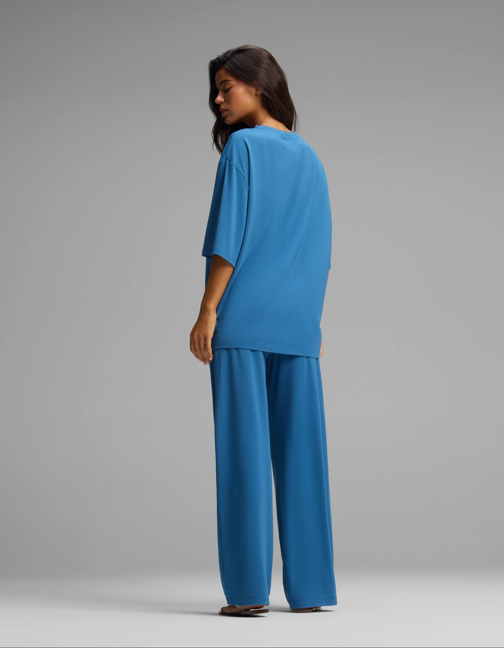 A woman with long dark hair, seen from behind, wears the Cozy Earth Women’s First Layer Boyfriend Sleep Tee & Pajama Pant Set in blue—a coordinated set featuring an oversized short-sleeve top and wide-leg pants—while posing against a solid grey background.