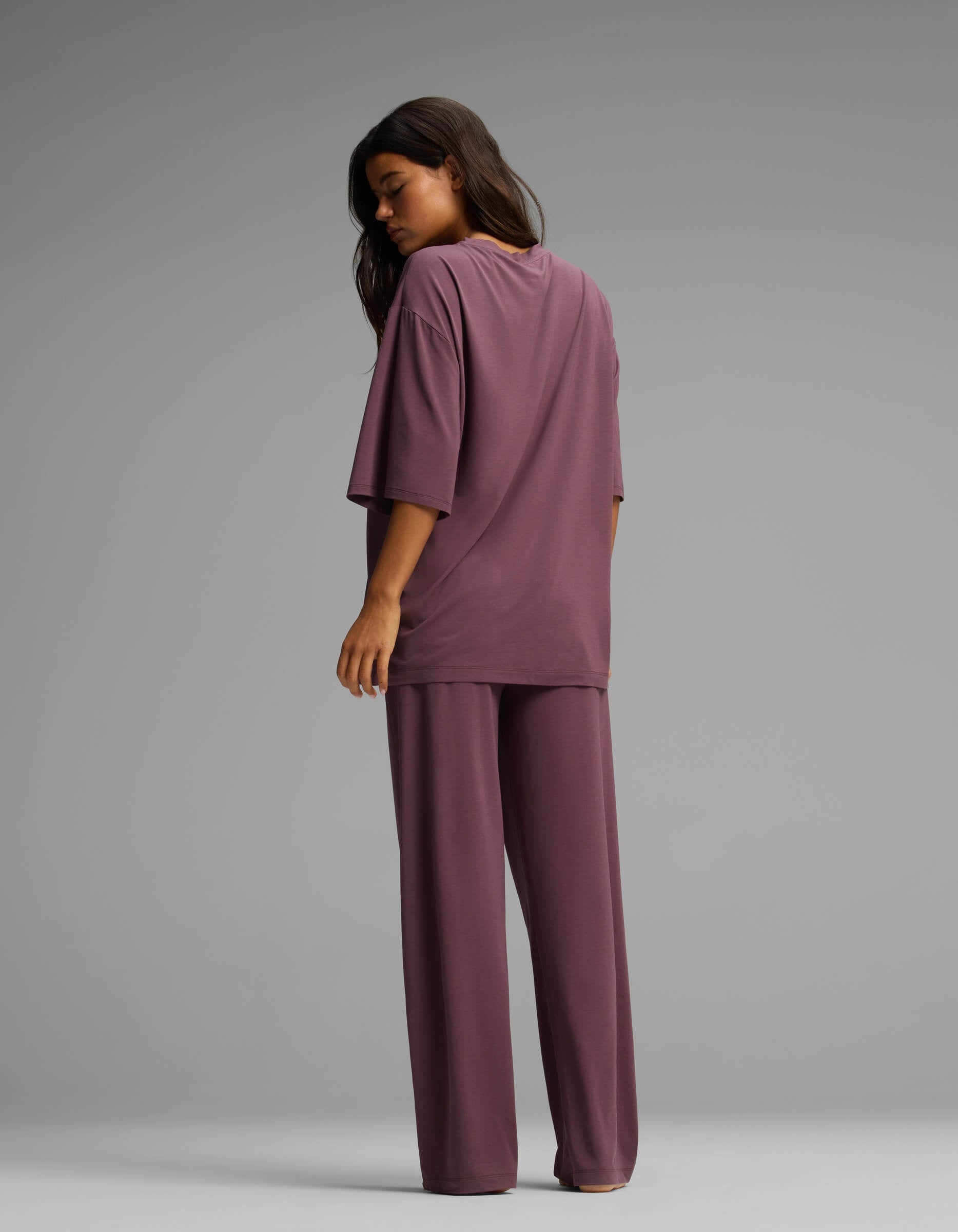 A woman stands facing away, wearing the Cozy Earth Women’s First Layer Boyfriend Sleep Tee & Pajama Pant Set—a loose-fitting, mauve-colored short-sleeve top and matching wide-legged pants—against a plain gray background.|Color:Fig